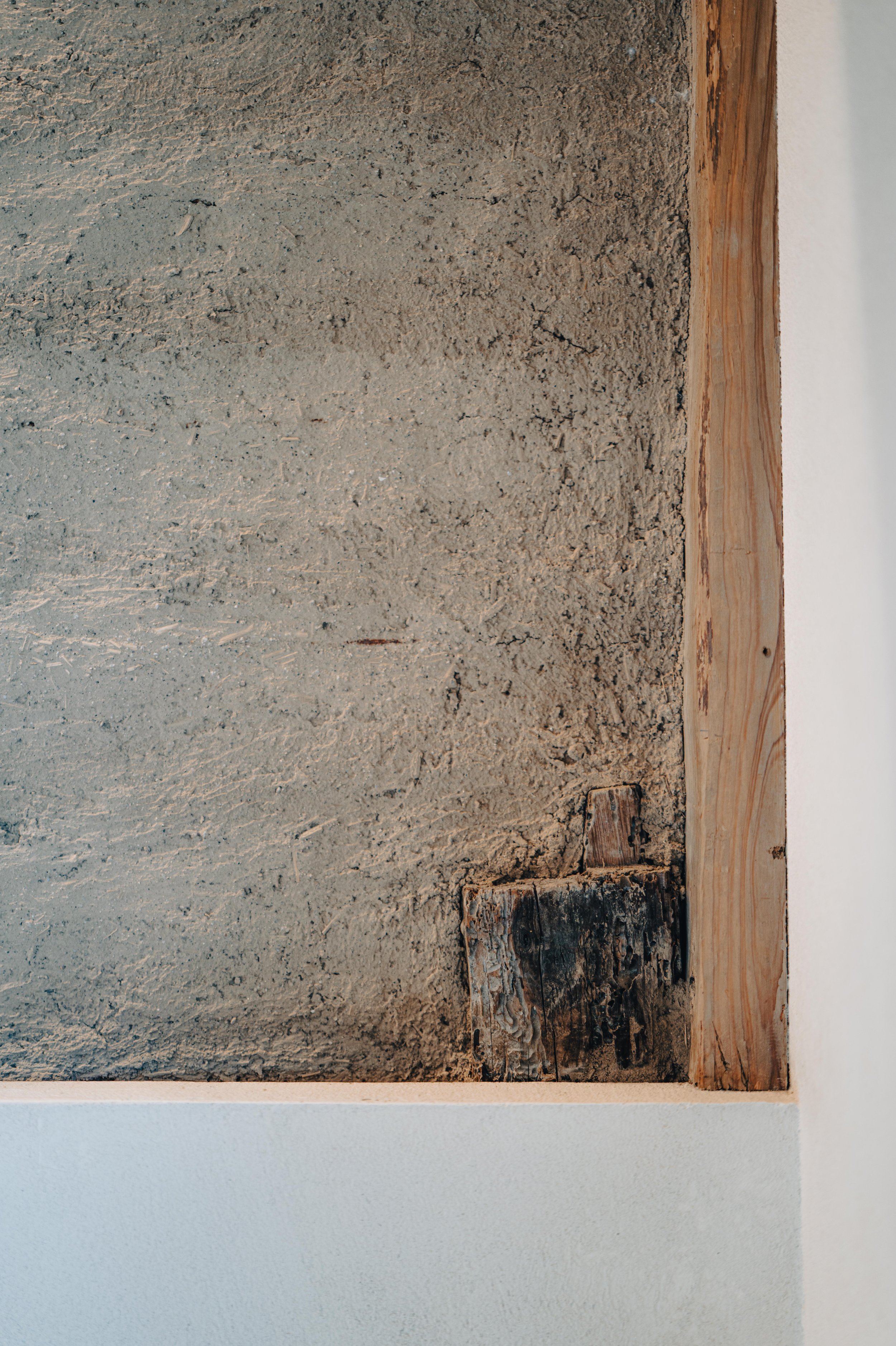 NUUT Aewol 눗애월 Close-up of a textured concrete wall with a wooden frame on the right side and a white painted surface at the bottom.
