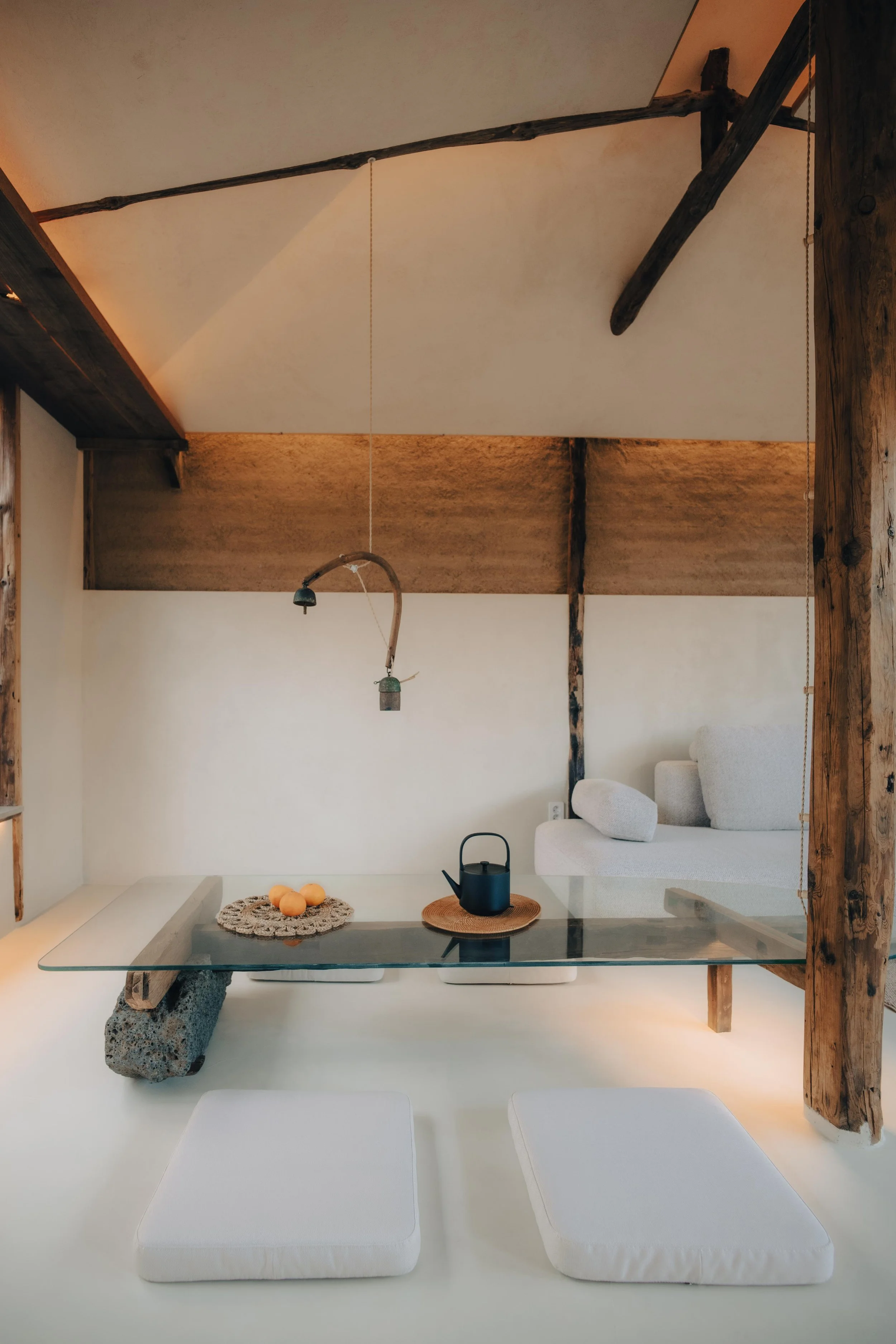 NUUT Aewol 눗애월 Minimalist room with a glass table on a large rock, two white floor cushions, a hanging lamp, a teapot on a woven mat, and a white couch in the background with wooden beams and a sloped ceiling.