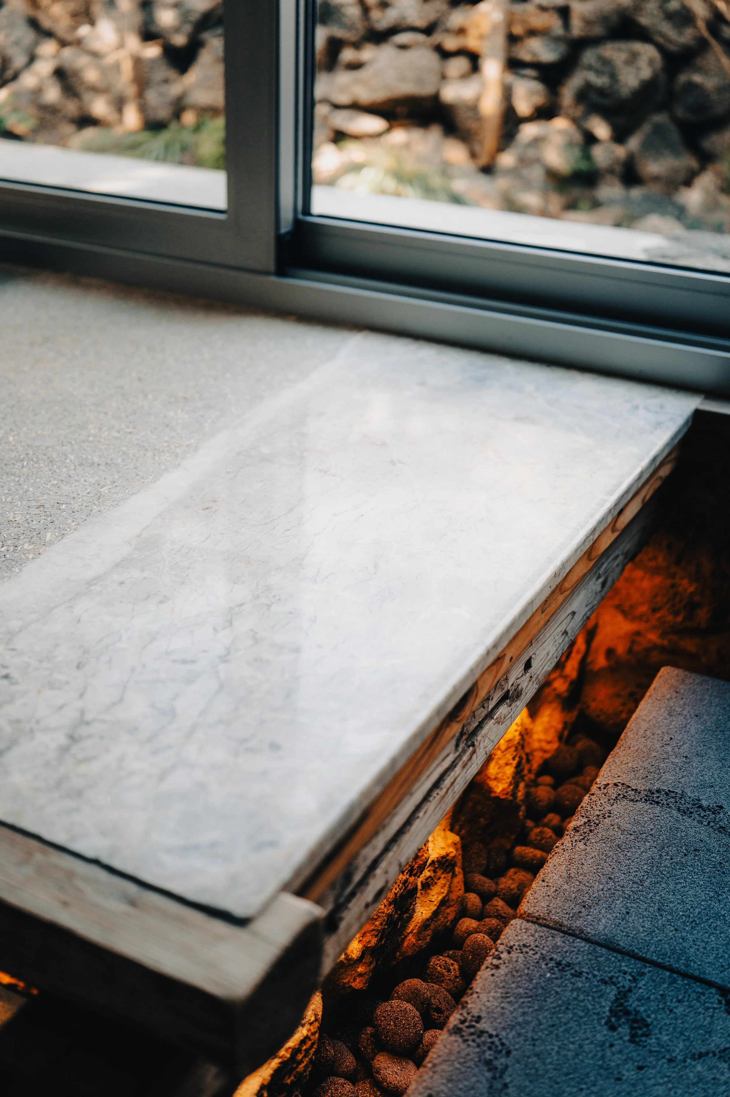 Close-up of a marble window sill with a sliding glass window and view of rocks outside, with warm lighting underneath the sill.