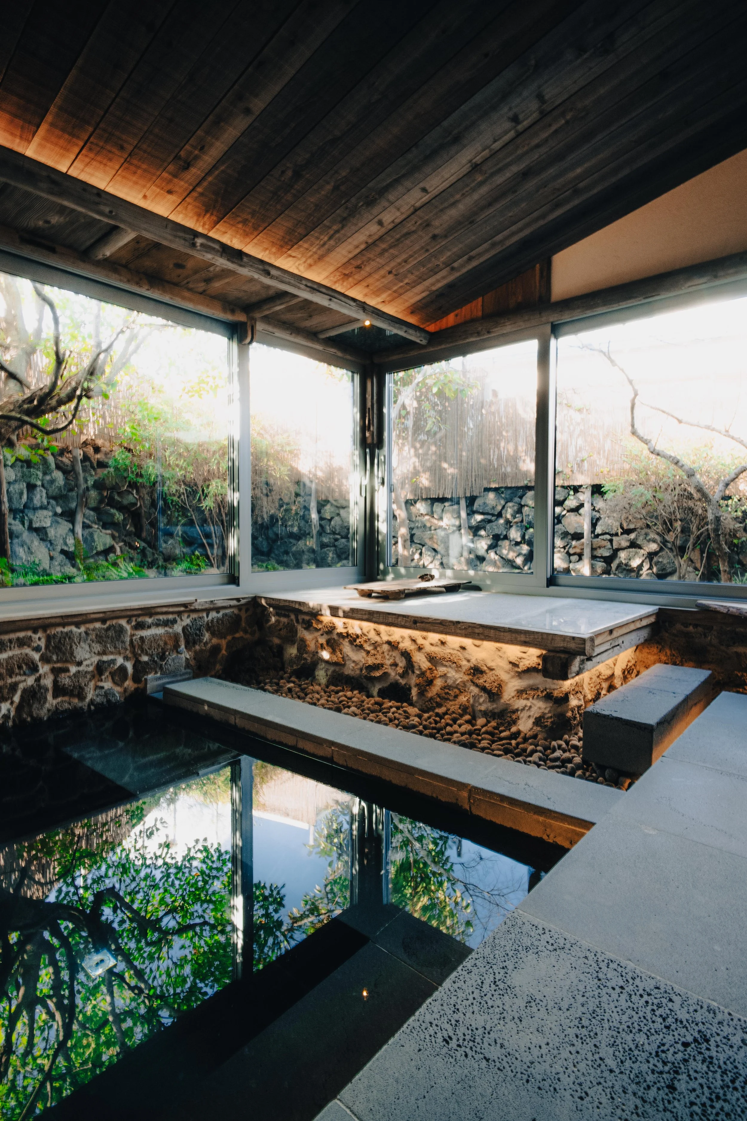 Indoor hot spring with black tile pool, surrounded by glass windows overlooking a garden with rocks, trees, and sunlight.