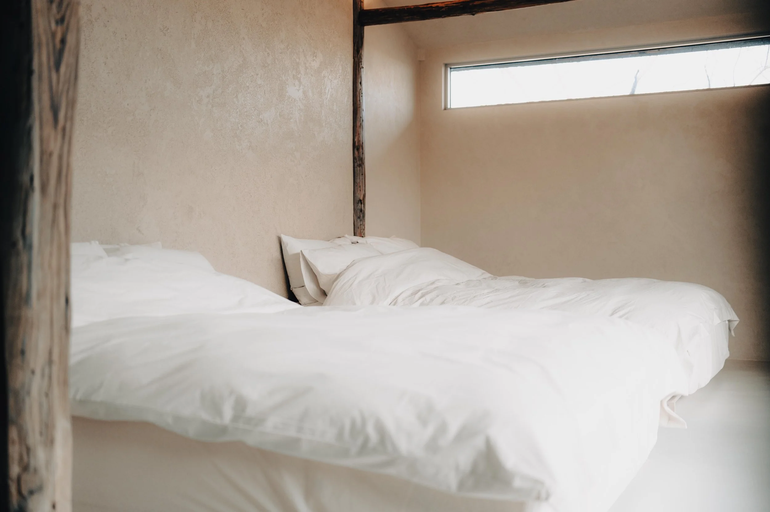 NUUT Aewol 눗애월 A minimalist bedroom with two unmade beds covered in white linens, beige textured walls, a narrow horizontal window, and exposed wooden beams.