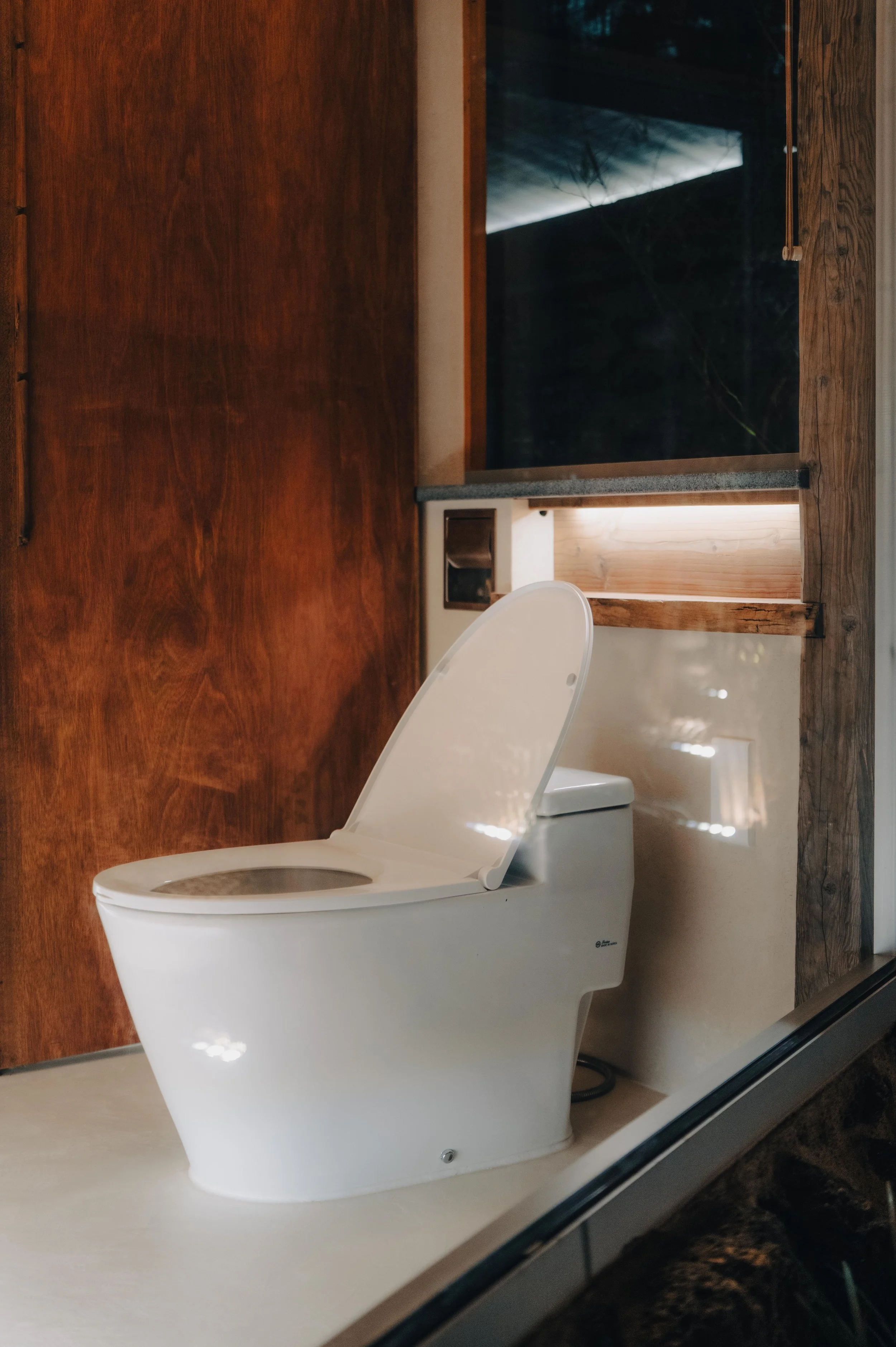 NUUT Aewol 눗애월 Modern white toilet with lid open in a rustic bathroom with wood paneling and a window.