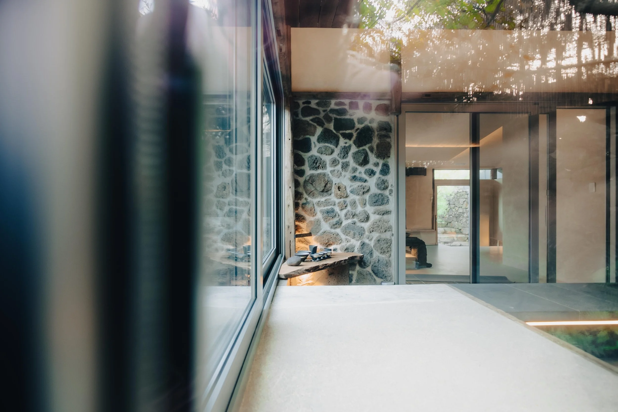 Interior view of a modern house with glass sliding doors, a stone wall, and a small wooden shelf with ceramic bowls, seen from an angle inside the room.