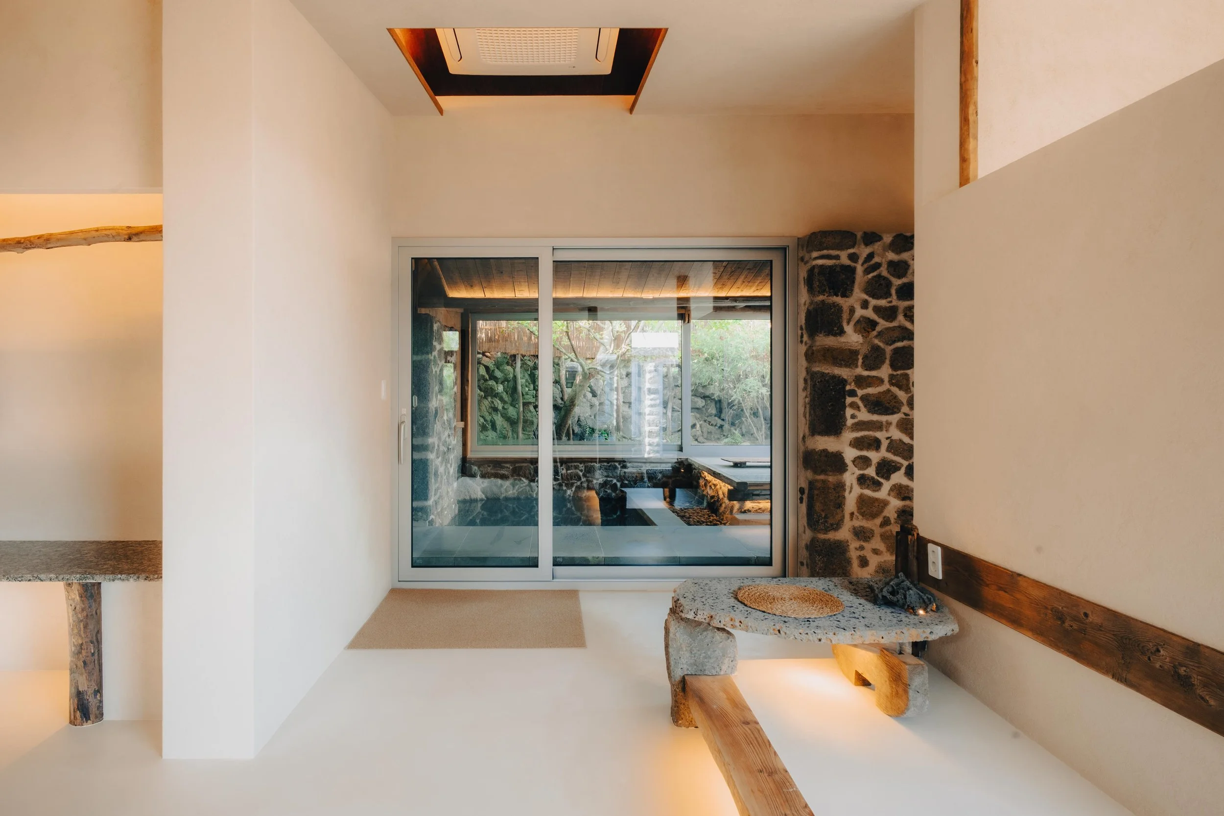 NUUT Aewol 눗애월 A minimalist interior room with sliding glass doors leading to a patio and garden, stone walls, a stone table with a woven mat, and wooden beams.