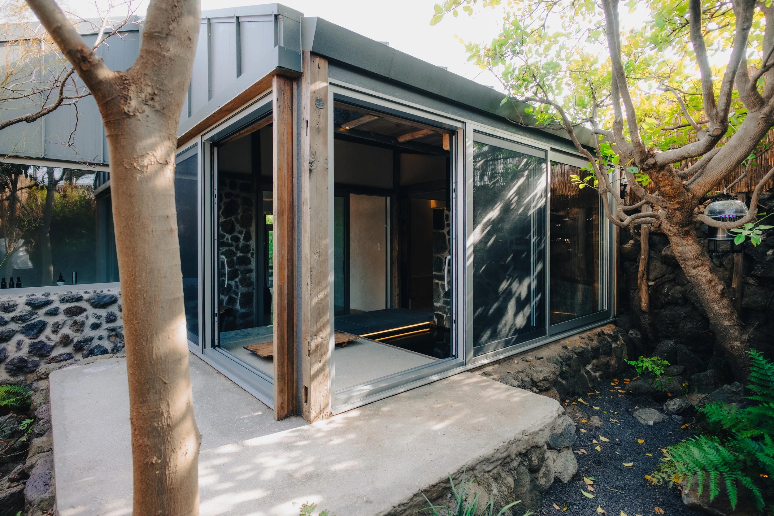 NUUT Aewol 눗애월 A modern tiny house with large glass sliding doors, surrounded by trees and rocks, with a concrete porch and natural wood accents.