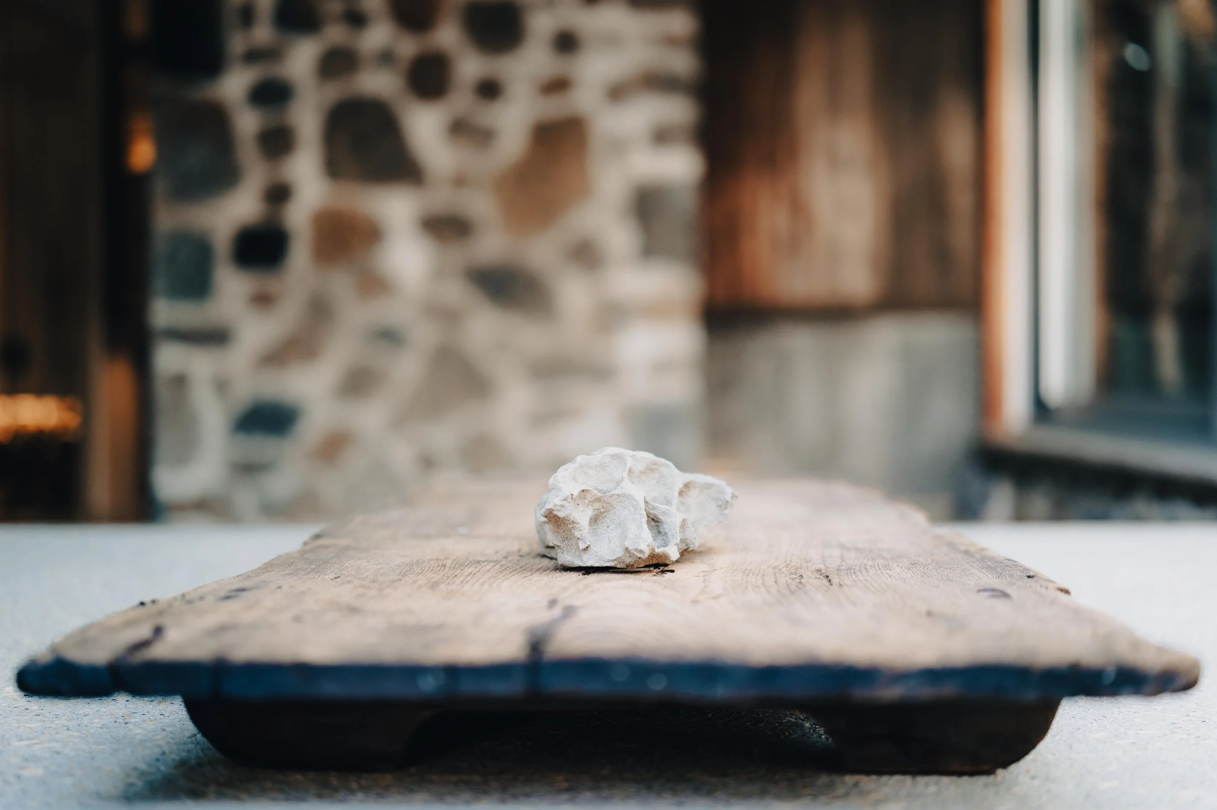 A small white rock resting on a wooden sushi board with a blurred stone wall and window in the background.