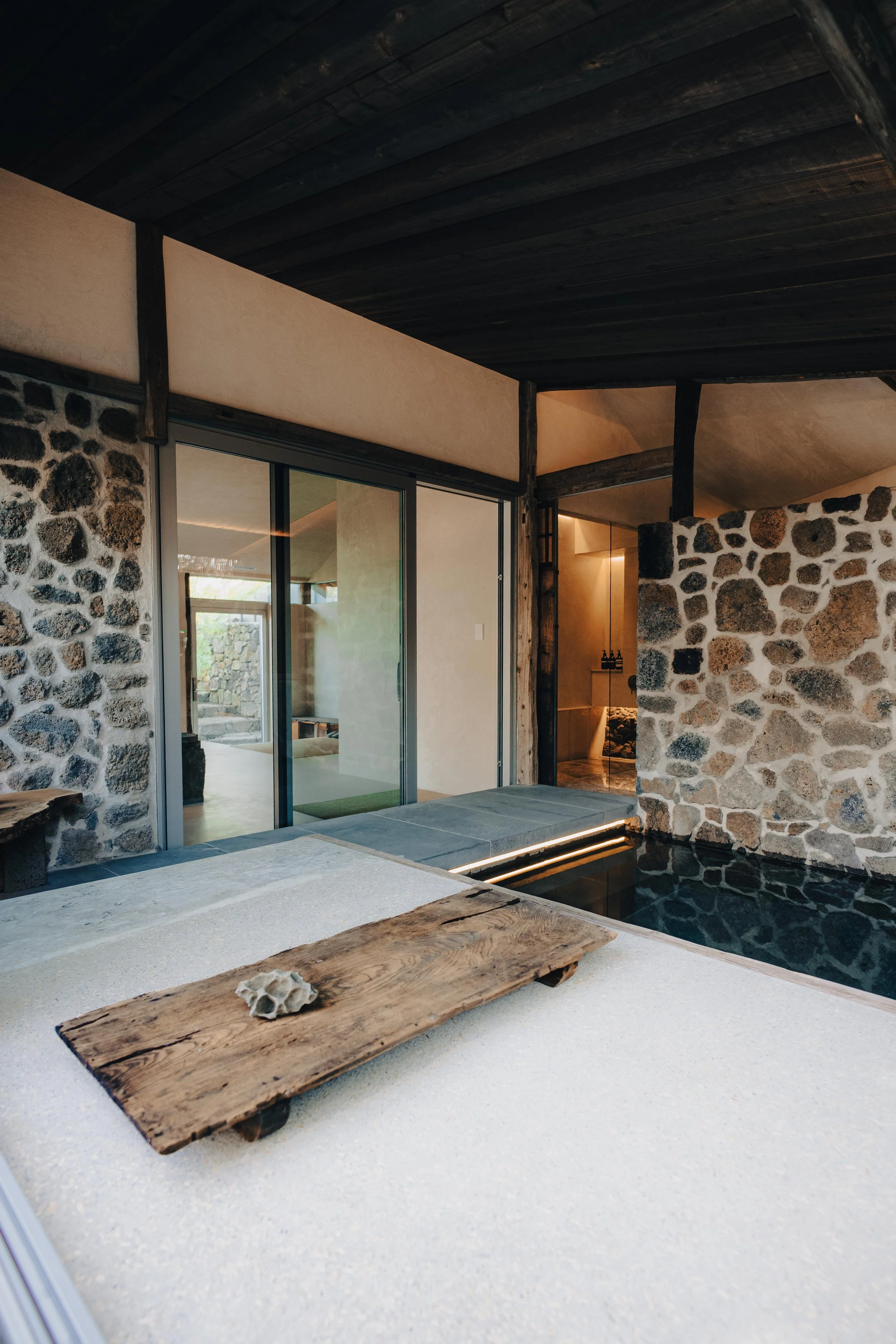 NUUT Aewol 눗애월 Modern indoor space with stone and wood accents, sliding glass doors, and a small pool or water feature.