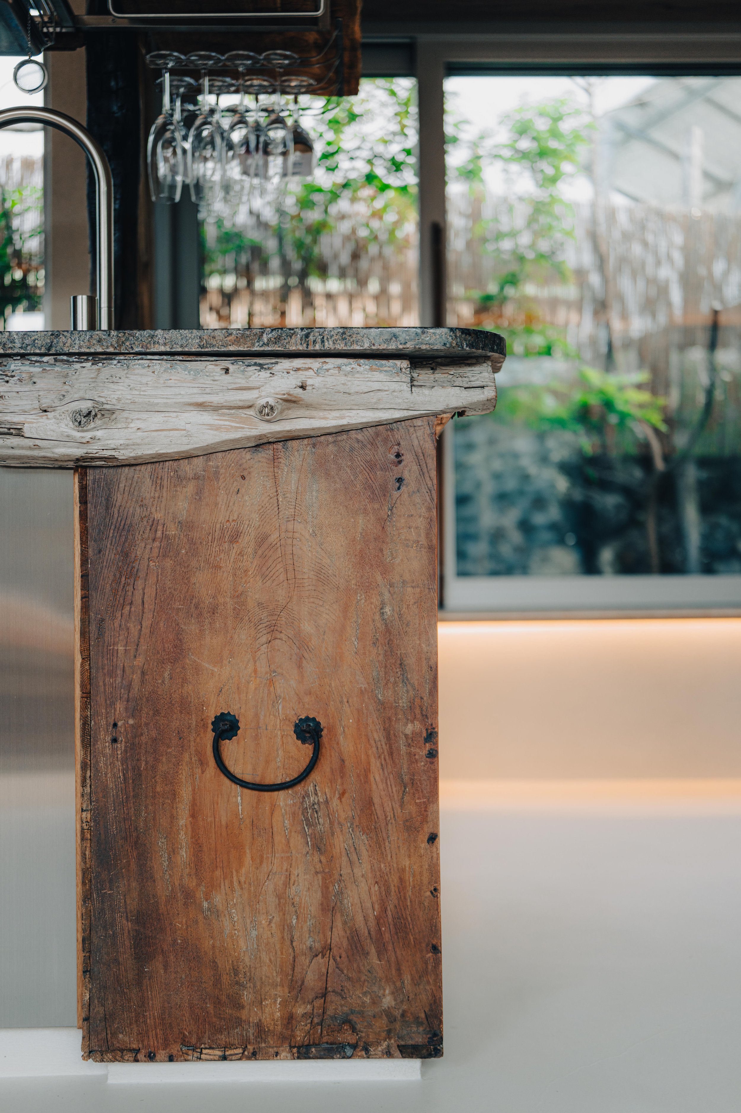 NUUT Aewol 눗애월 Close-up of a rustic wooden kitchen cabinet with a smiley face metal handle, countertop, and hanging wine glasses in the background.