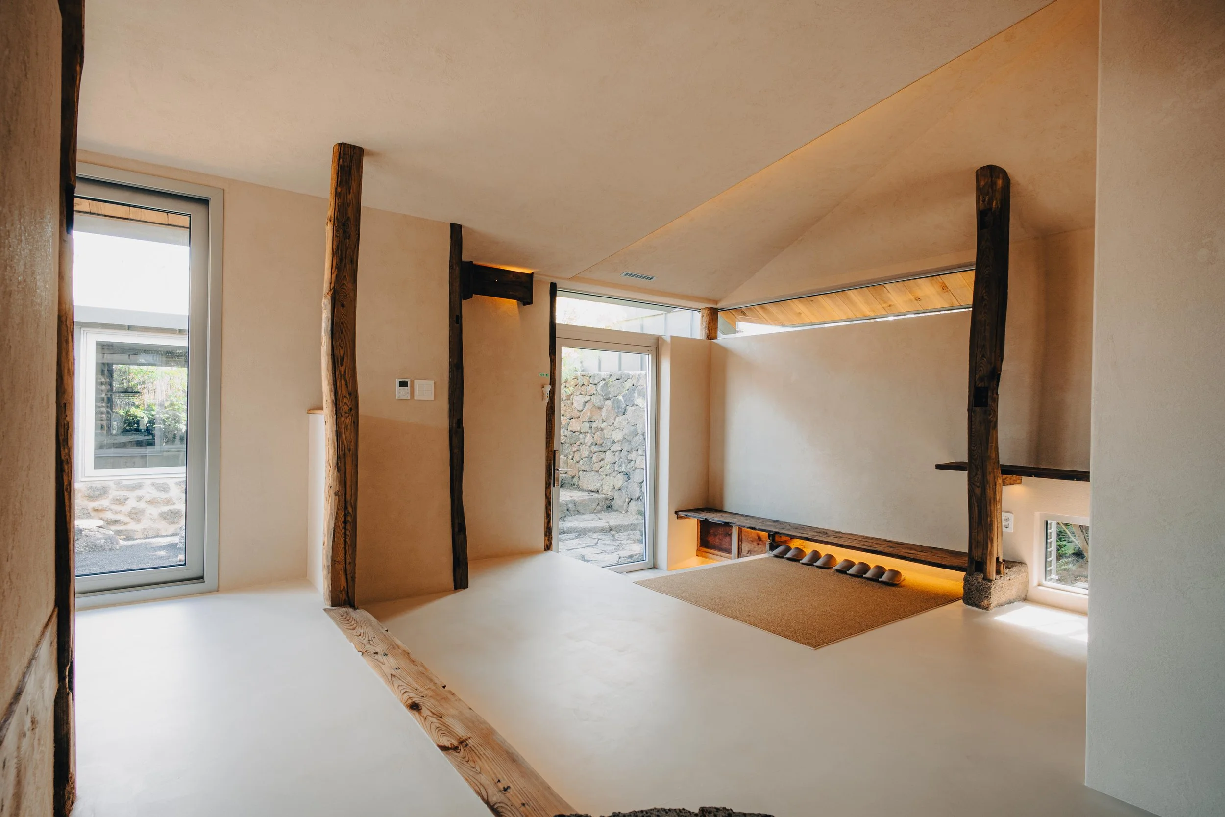 NUUT Aewol 눗애월 Interior view of a modern house with beige walls, wooden beams, a glass door leading outside, a small window, and minimalistic decor.
