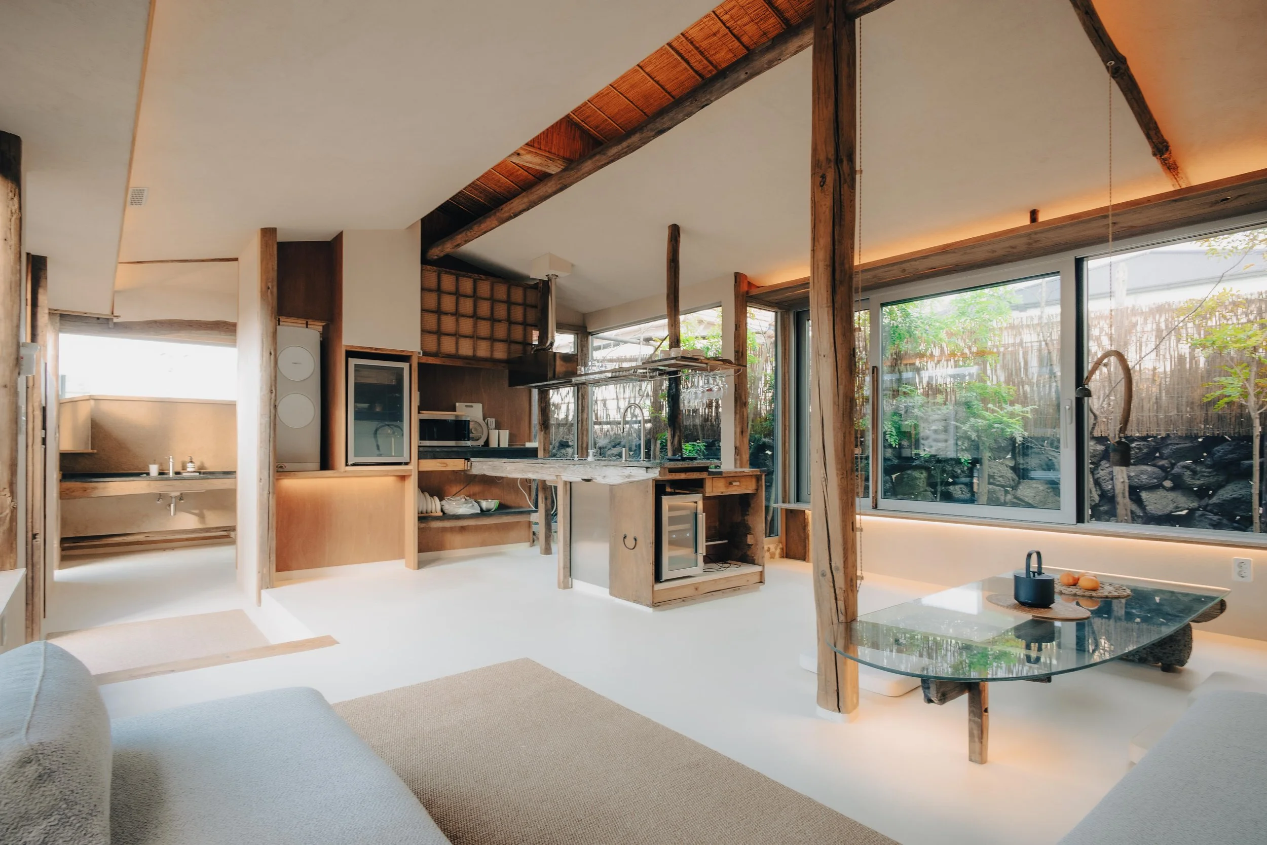 NUUT Aewol 눗애월 Interior of a modern, rustic-style living space with large windows, wooden beams, a kitchen area, and a seating area with a glass table.