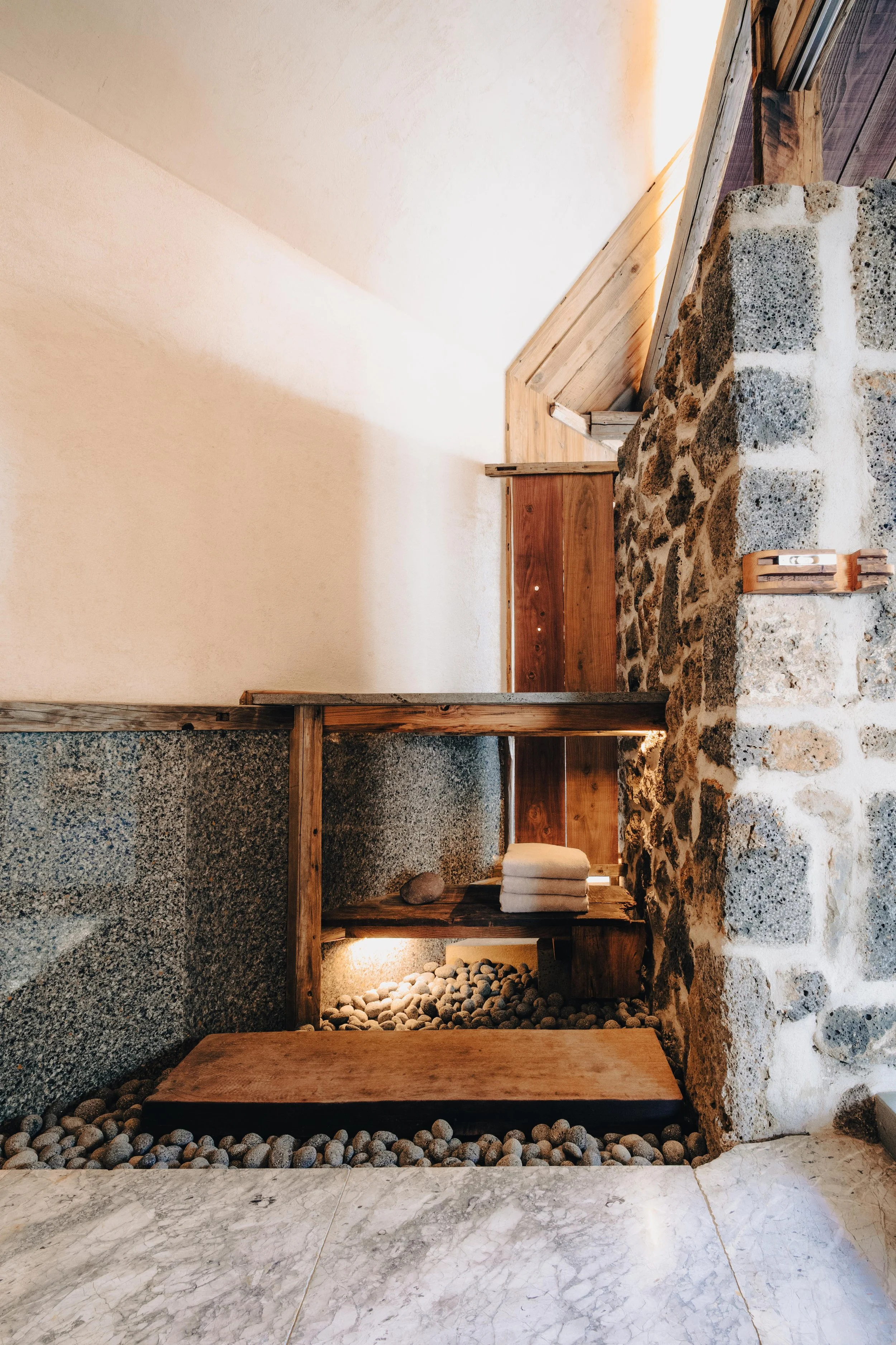 NUUT Aewol 눗애월 Interior of a rustic spa or sauna with a stone wall, wooden benches, and towels, illuminated by soft lighting.