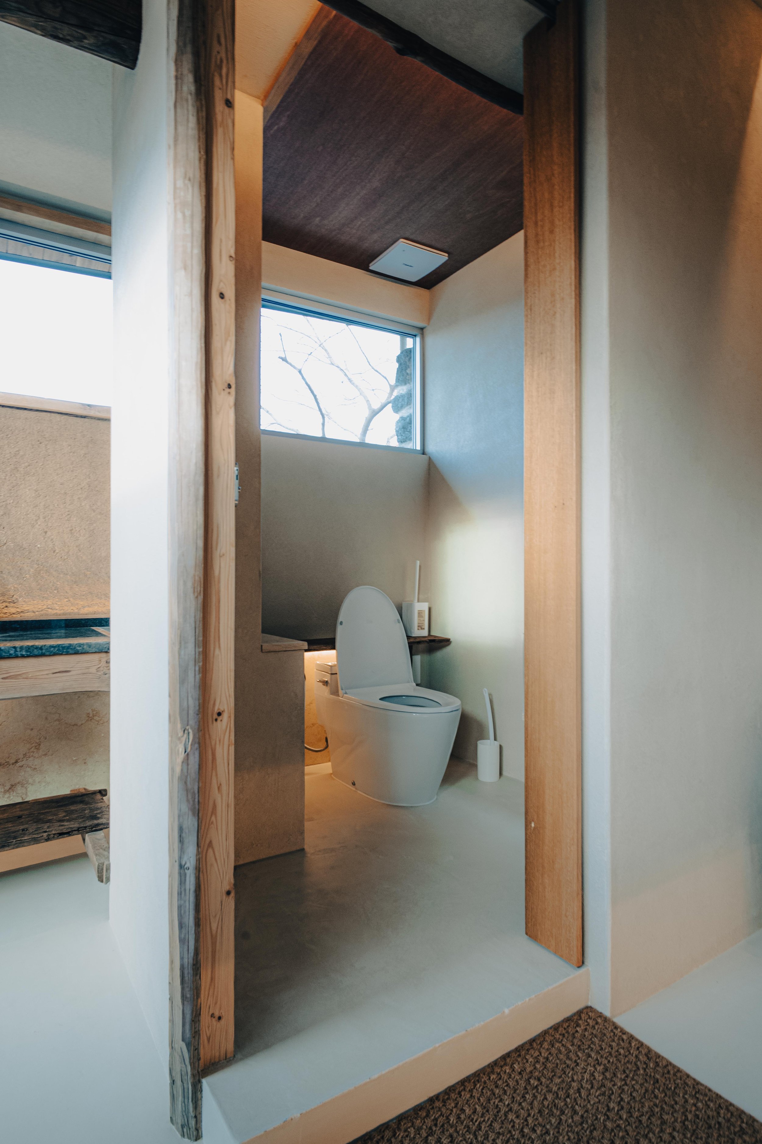 NUUT Aewol 눗애월 A modern bathroom featuring a white toilet with an open lid, natural light coming through a large window, and minimalist decor with a toilet brush on the floor.