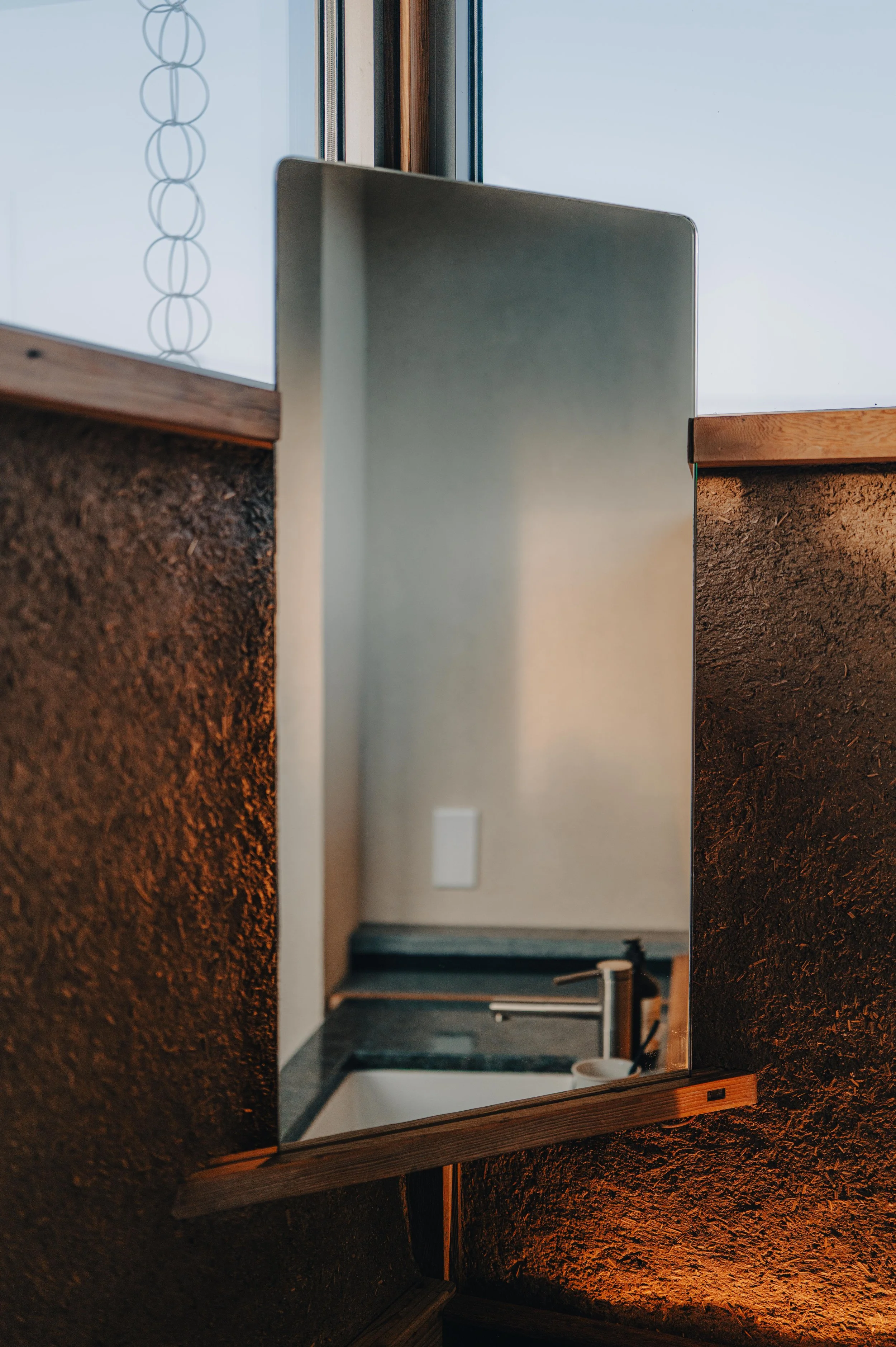 NUUT Aewol 눗애월 A mirror mounted on a textured, dark brown wall, reflecting a sinks and faucet area with a window above.