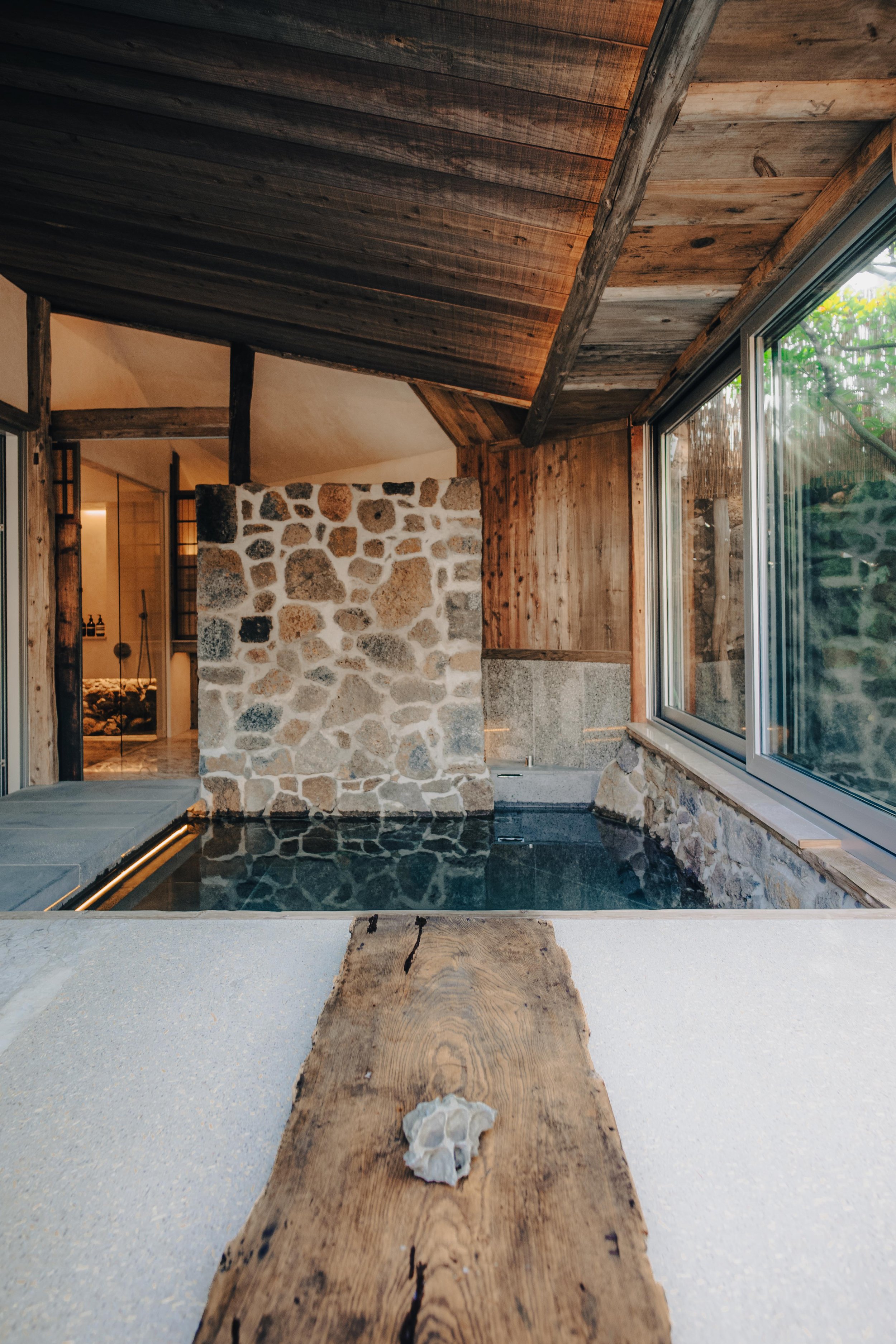 NUUT Aewol 눗애월 Indoor pool with stone walls, wooden ceiling, and large windows in a rustic style.