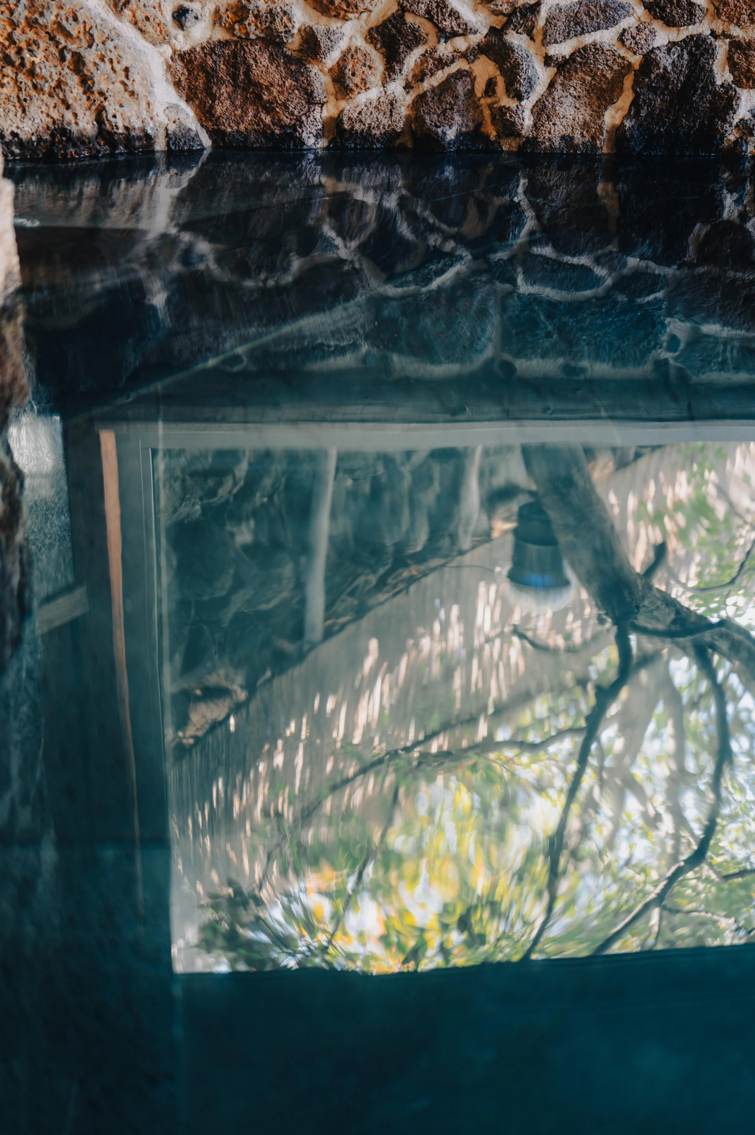 A close-up view of a water feature with rocks and a glass panel, reflecting trees and sky.