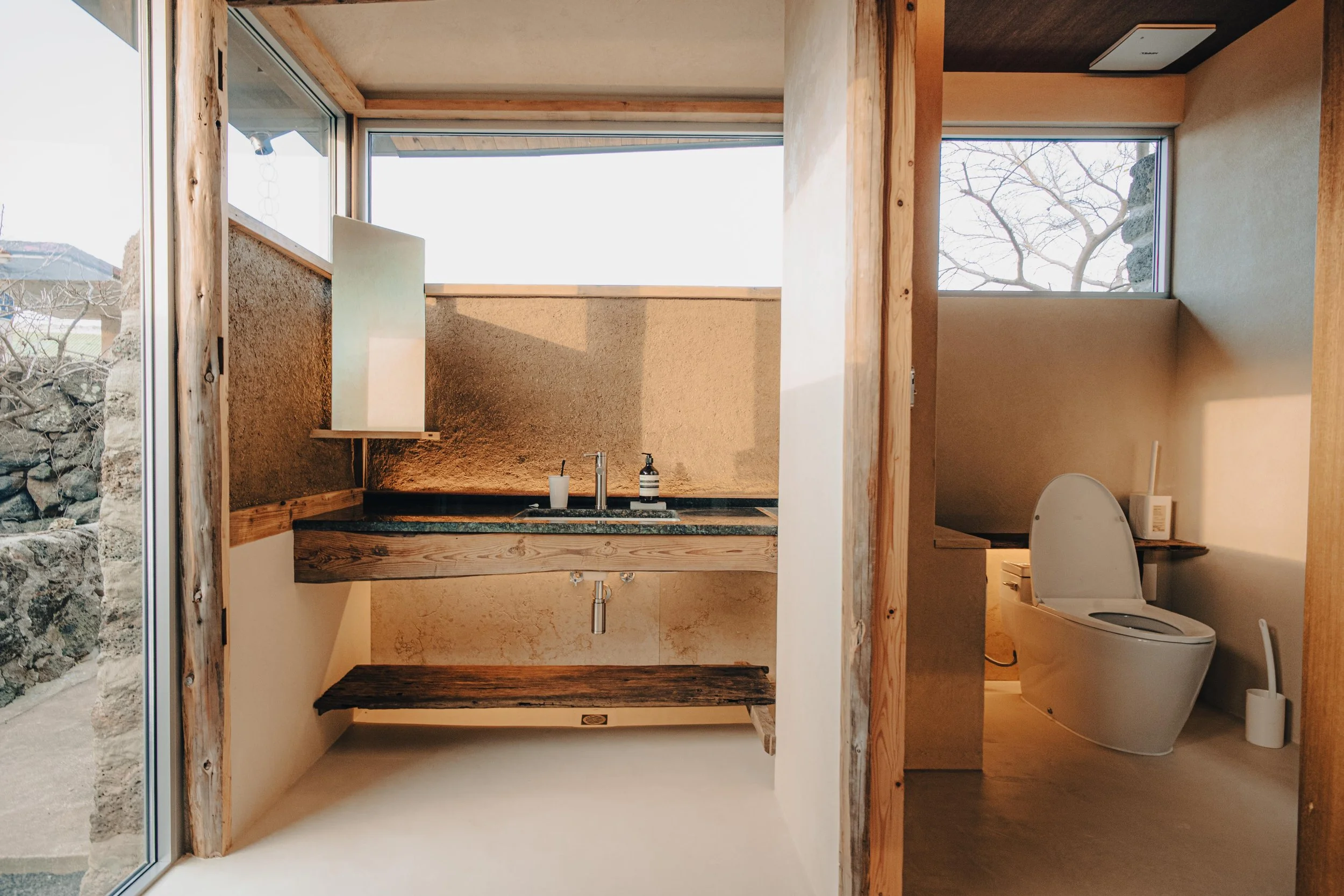NUUT Aewol 눗애월 Bathroom with a rustic wooden vanity, black countertop, faucet, soap dispenser, and a white cup, separated from the toilet area by a wall, with natural light coming through large windows.
