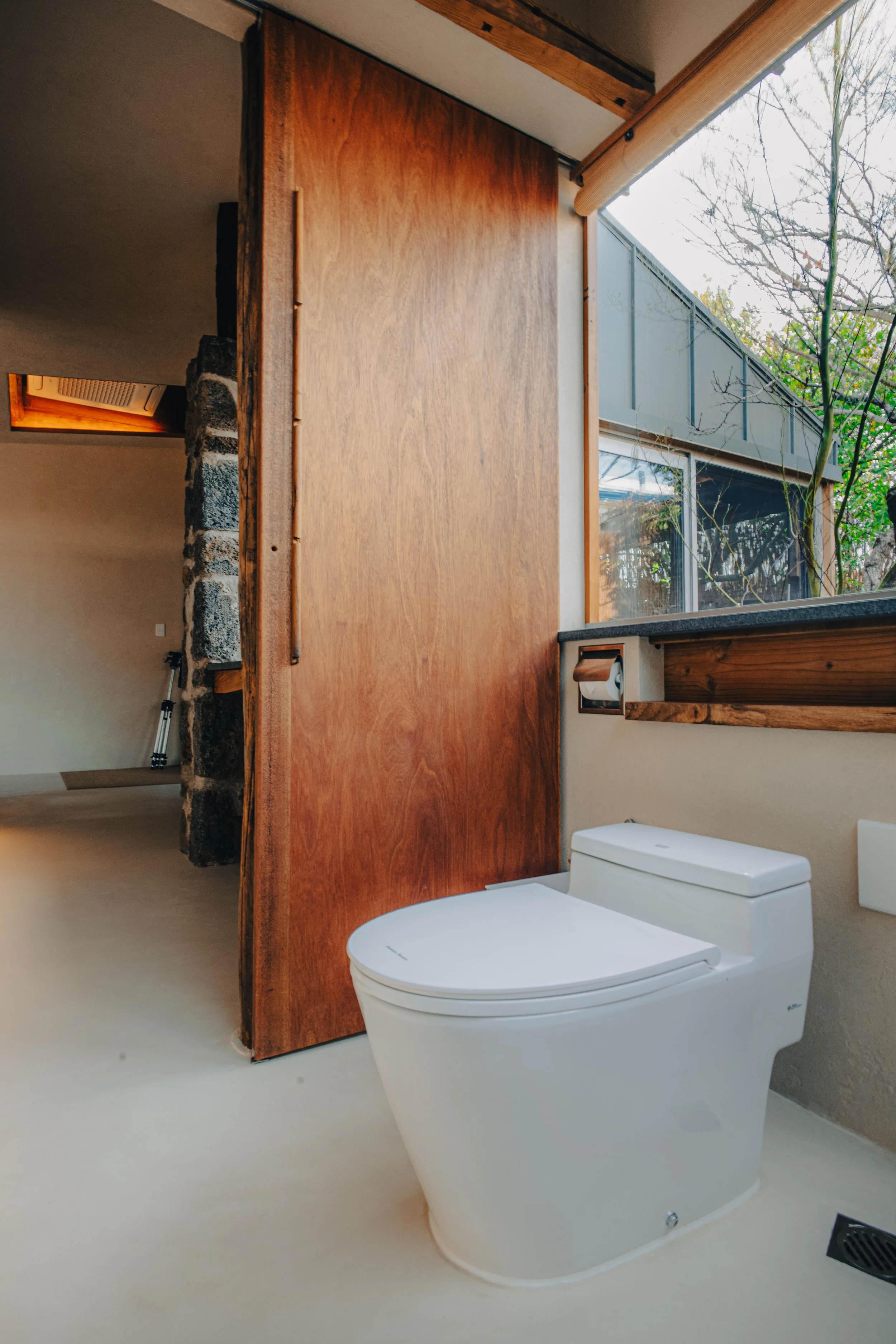 NUUT Aewol 눗애월Modern bathroom with a white advanced toilet, wooden accents, large window with an outdoor view, and a stone wall section.