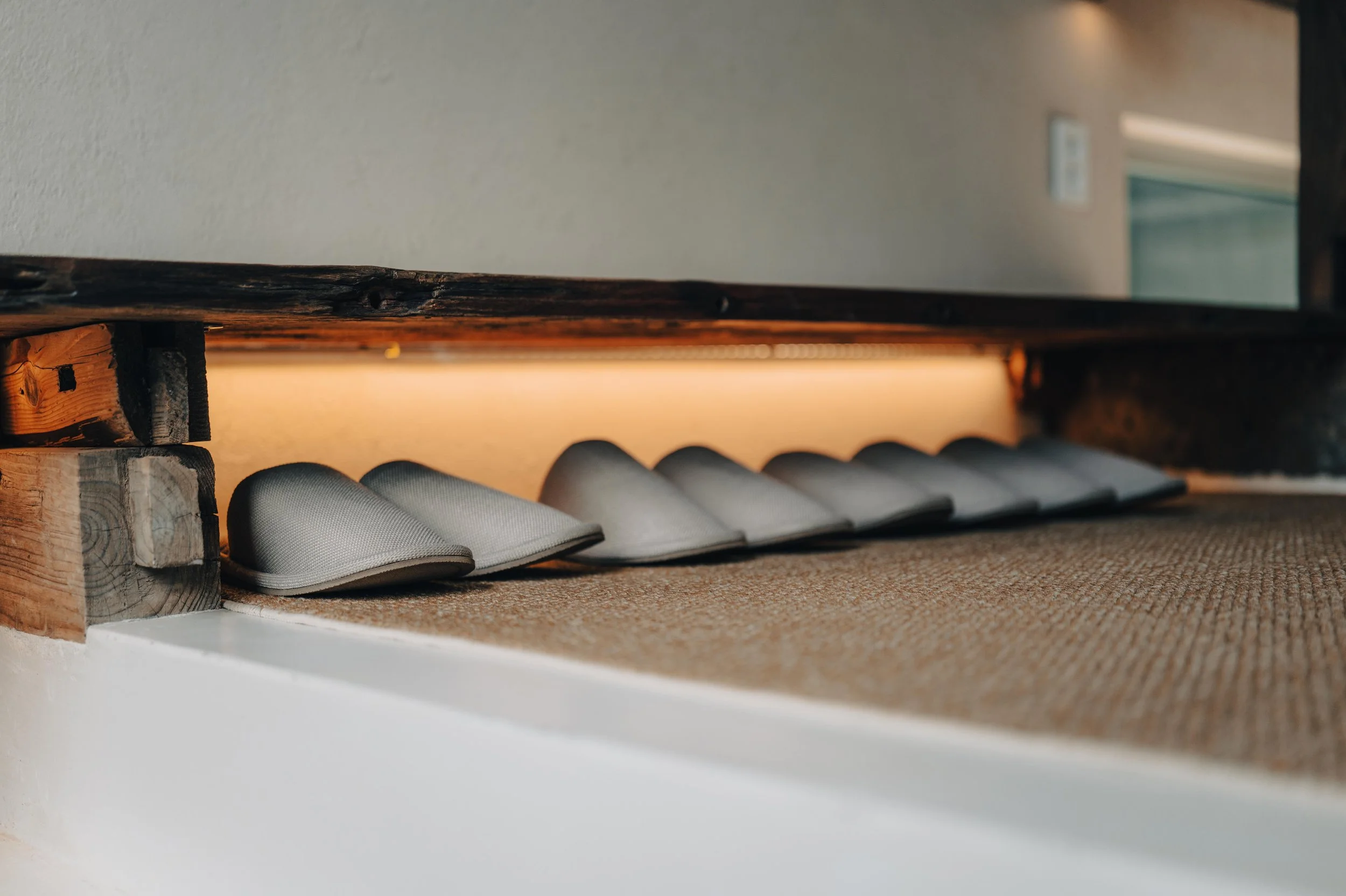 NUUT Aewol 눗애월 A row of gray slippers under a wooden ledge with warm lighting, on a beige textured mat, in an indoor setting.