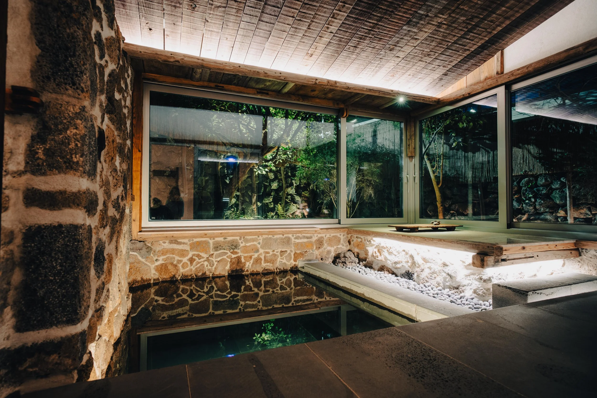 NUUT Aewol 눗애월 Interior of a rustic room with stone walls, large glass windows, and a view of a garden with trees. There is a small pond or water feature with white pebbles and a ledge with a tray on it. The ceiling is wooden with warm lighting.