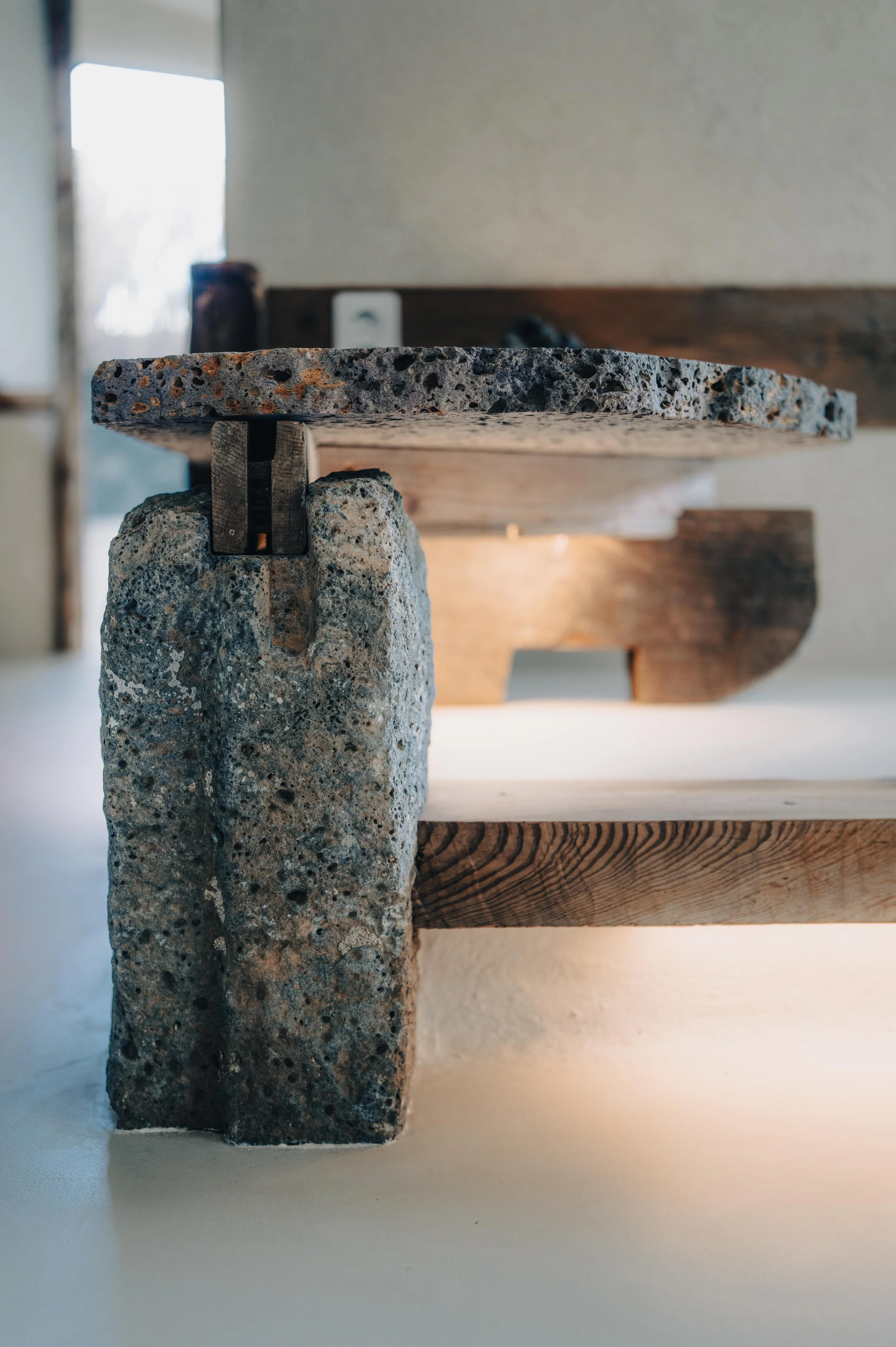 NUUT Aewol 눗애월 Close-up of a table leg made from a rough, gray, porous stone supporting a wooden tabletop with light-colored wood grain. The background shows a blurred interior scene with a window allowing natural light.