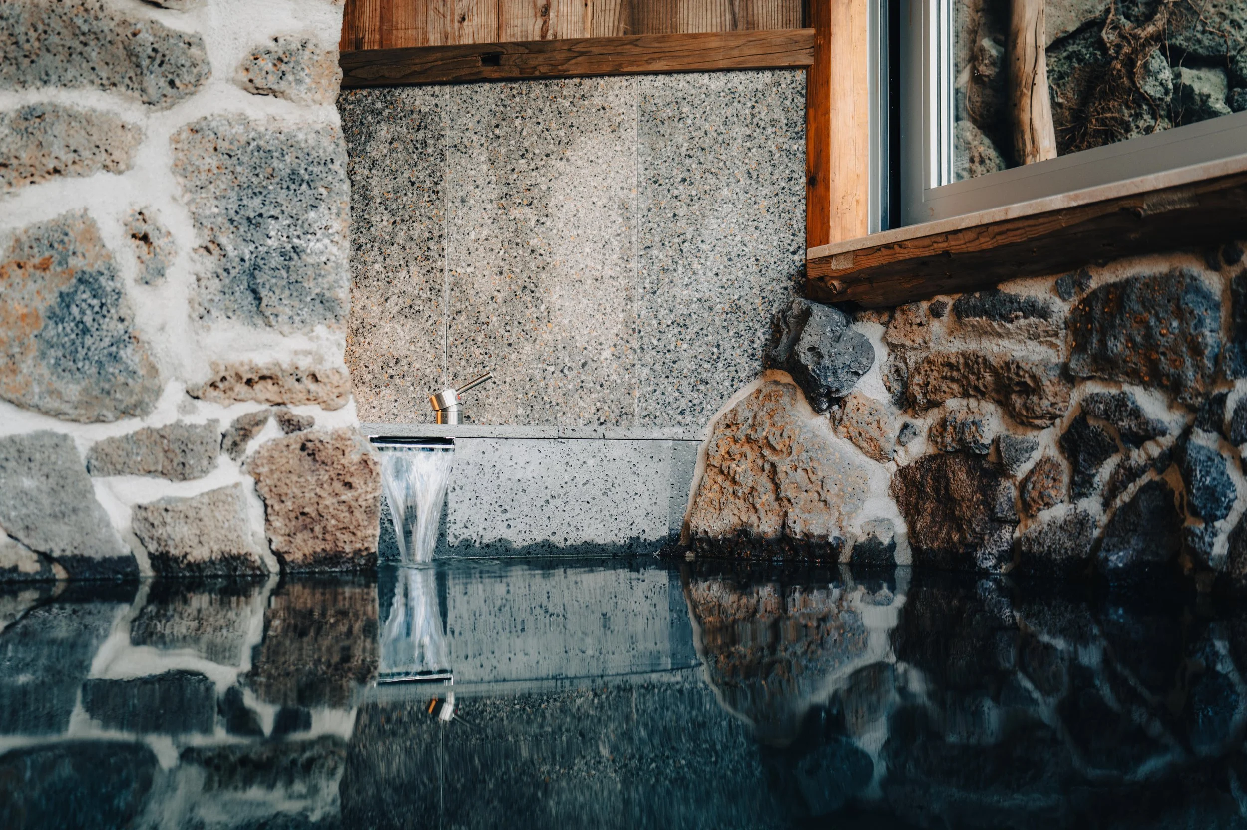 An indoor stone wall with a small fountain, decorated with large rocks and a window showing outside wooden logs.