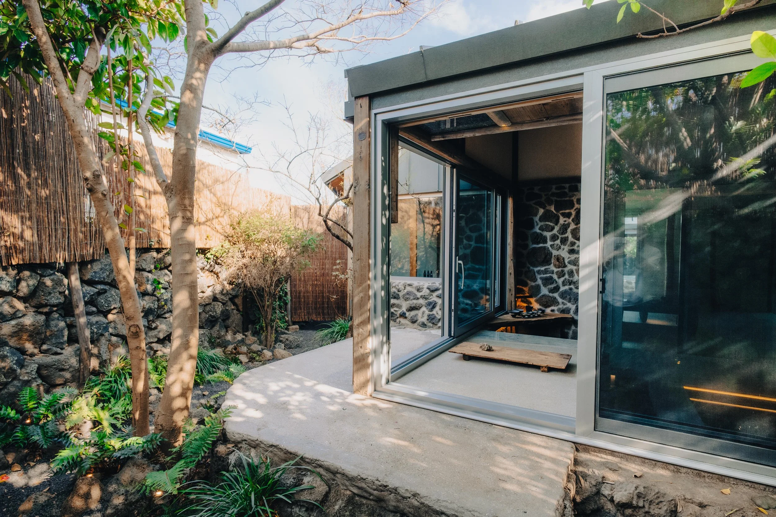 NUUT Aewol 눗애월 A modern outdoor kitchen with stone walls and glass sliding doors, surrounded by a garden with trees, rocks, and various plants.