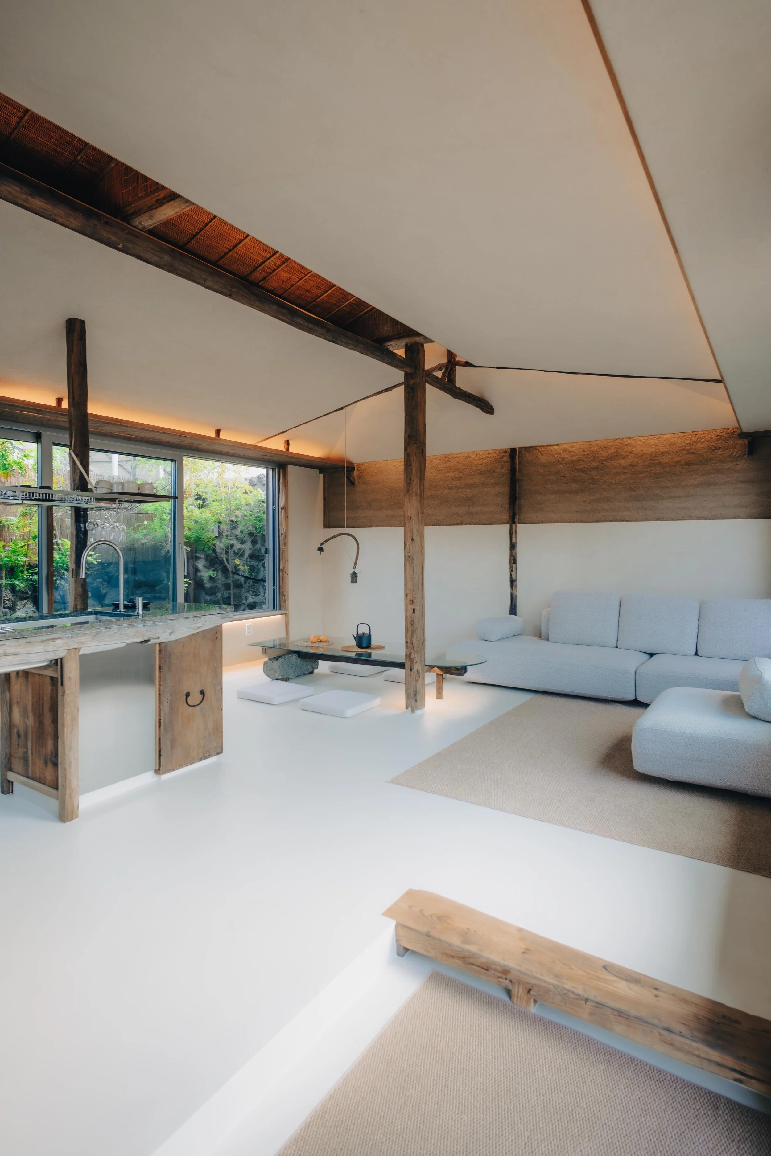 NUUT Aewol 눗애월 A minimalist living room with white walls, a light grey sofa, and wooden beams and accents, with a large window revealing greenery outside.