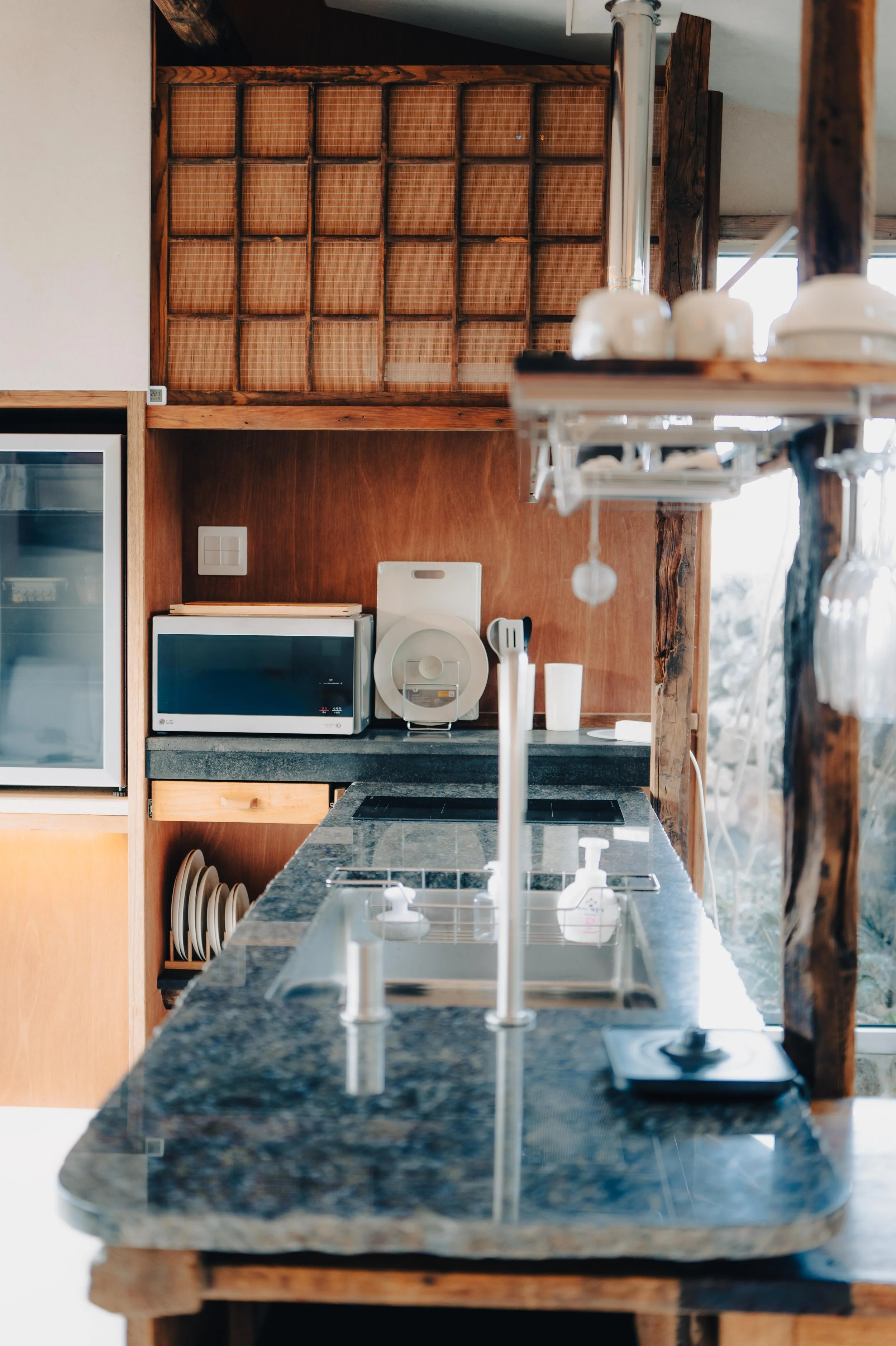 NUUT Aewol 눗애월 Kitchen with black granite countertop, sink, microwave, and various small appliances. Wooden shelves and cabinets, some with plates and mugs, and sunlight streaming through a window.