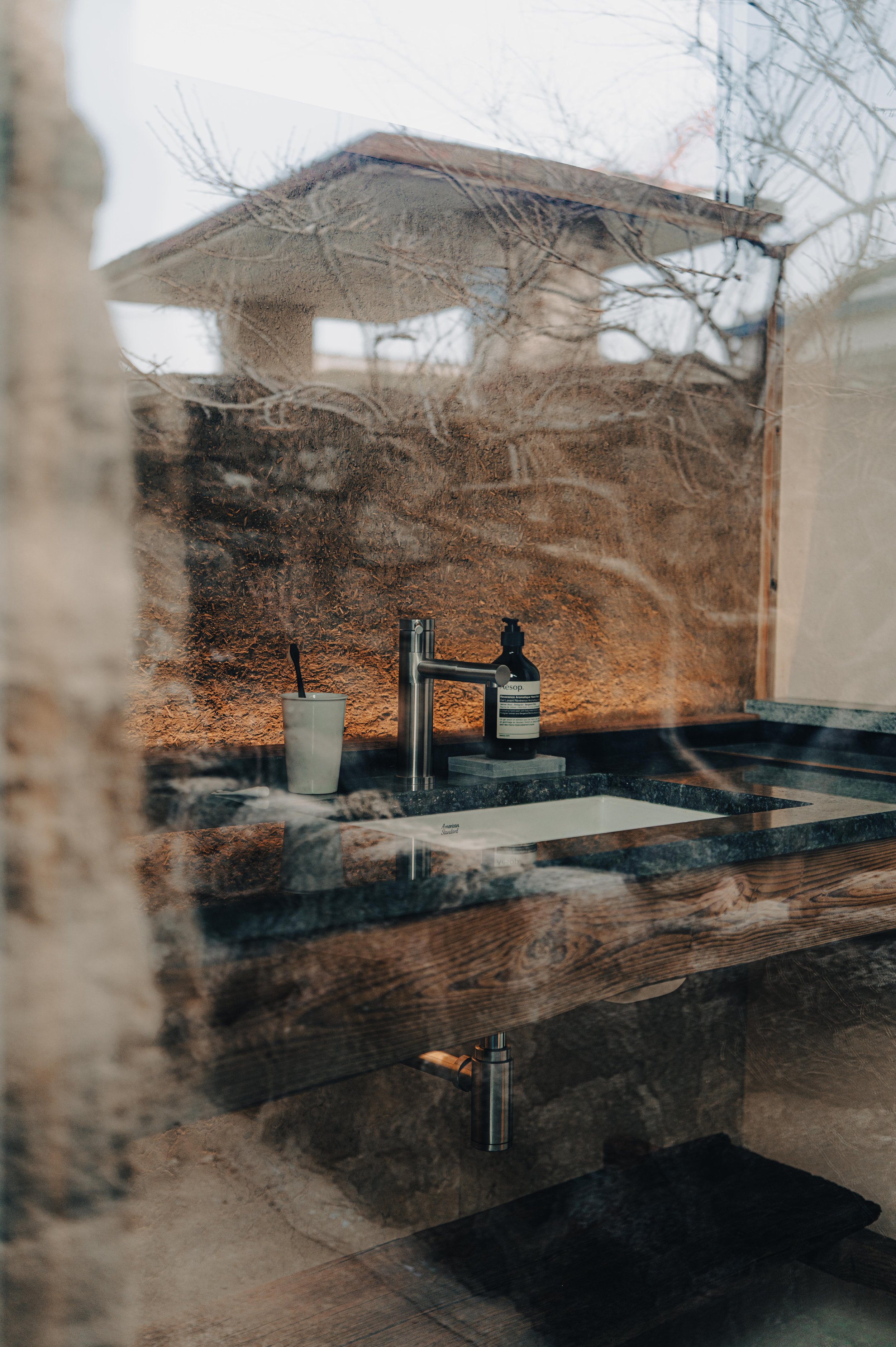 NUUT Aewol 눗애월 Indoor bathroom sink with a soap dispenser and a toothbrush in a cup, viewed through a glass with reflections of trees outside.