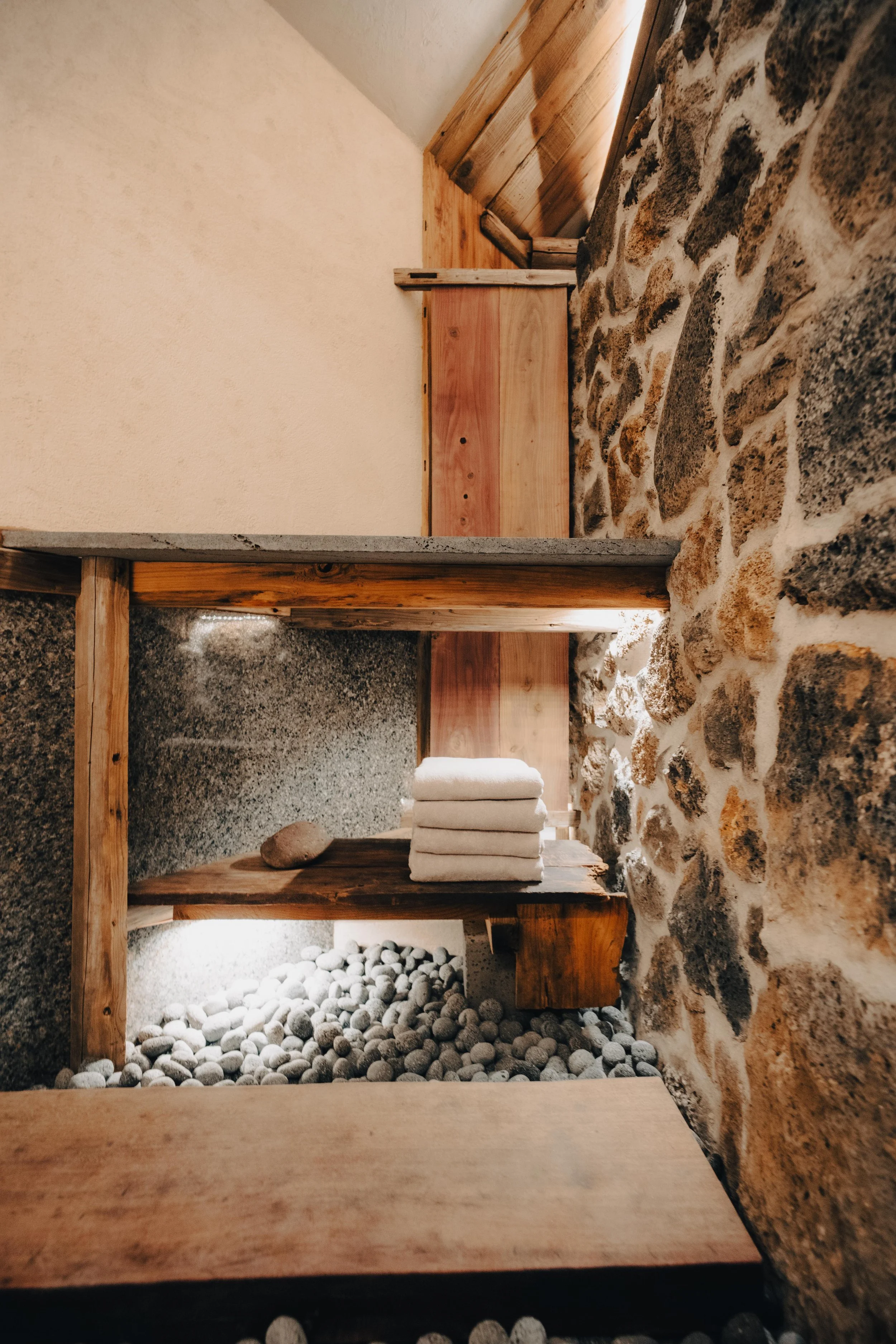 NUUT Aewol 눗애월 Spa or relaxation room with curved stone wall, wooden shelves, folded towels, and a pebble floor.