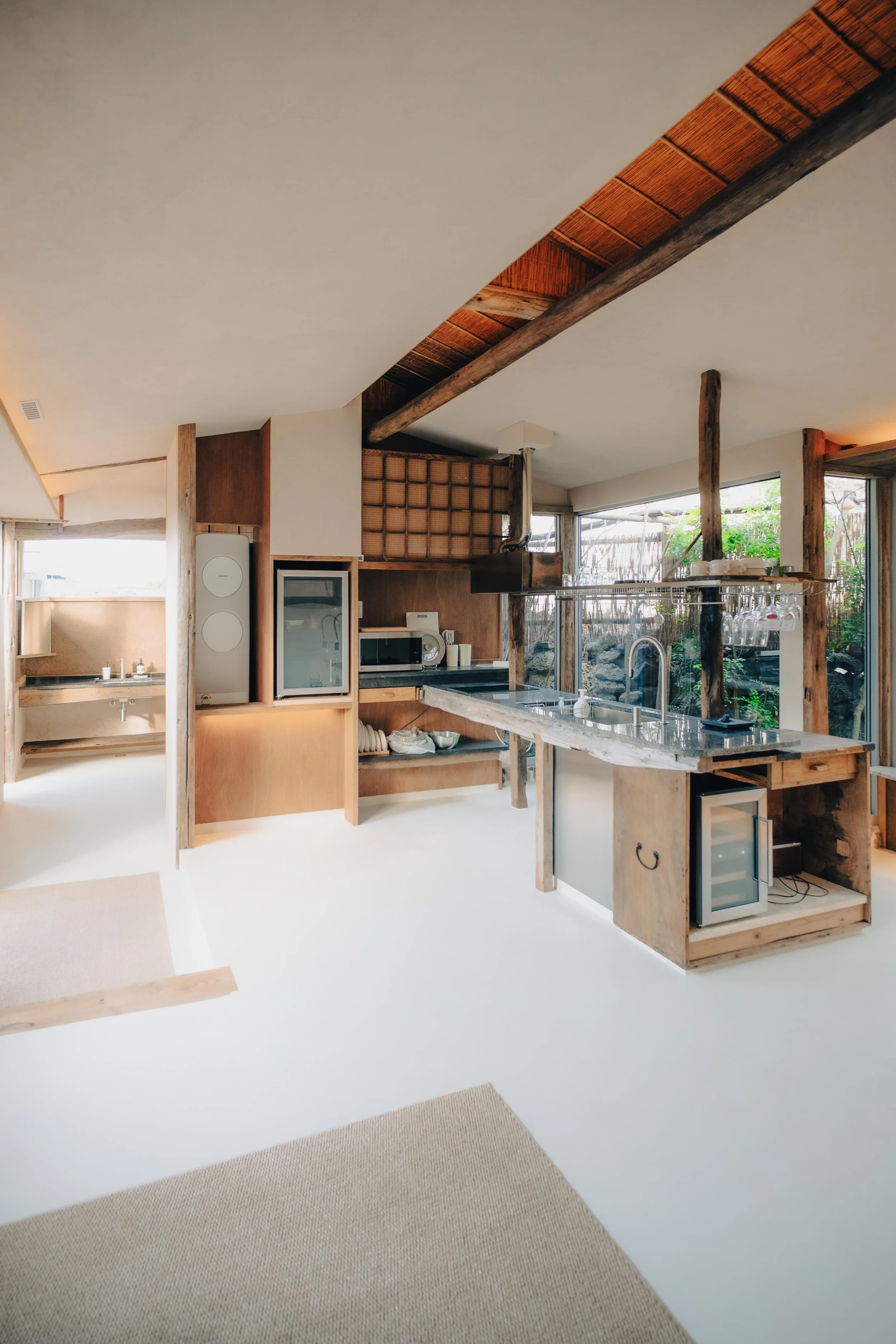 NUUT Aewol 눗애월Interior of a rustic modern kitchen with wooden beams, a small wine refrigerator, and sliding glass doors leading outside.