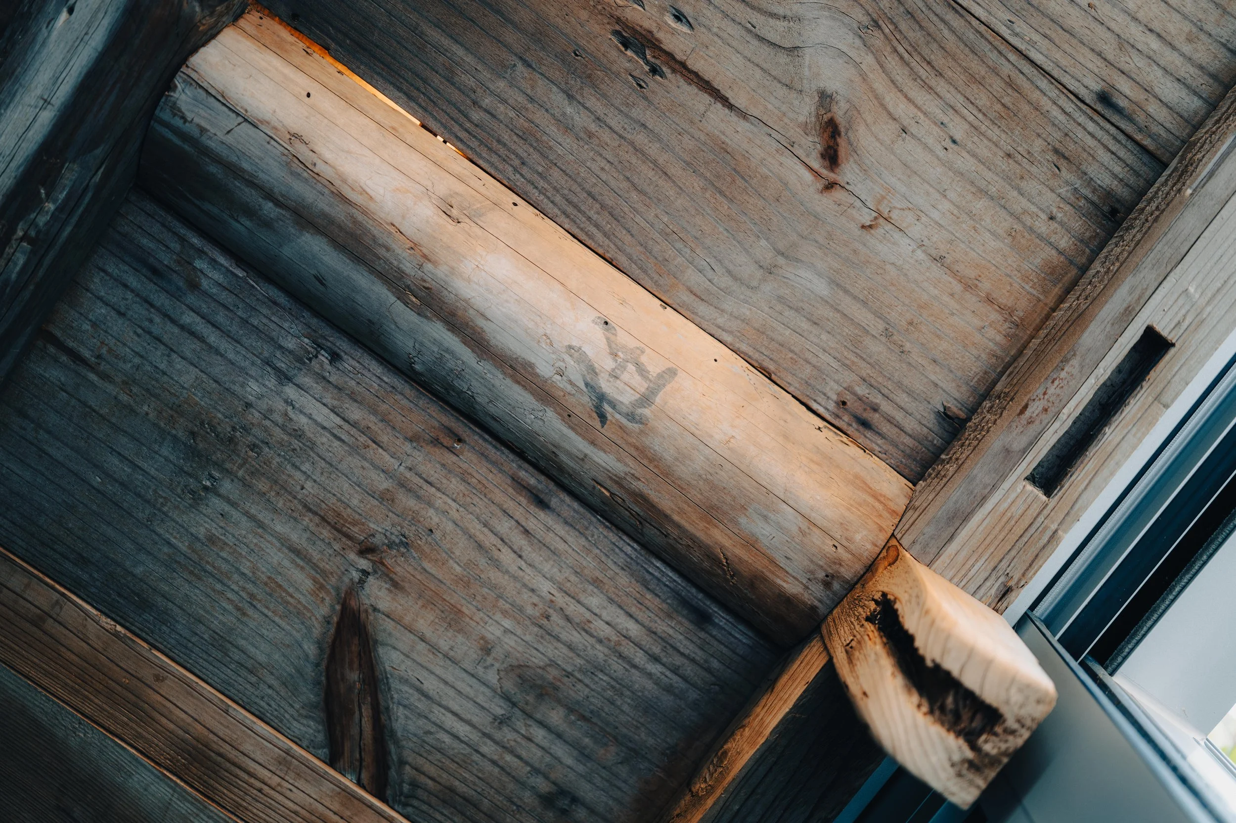 Close-up of a wooden corner with weathered wood and a window frame on the right side, showing the edge of the window.