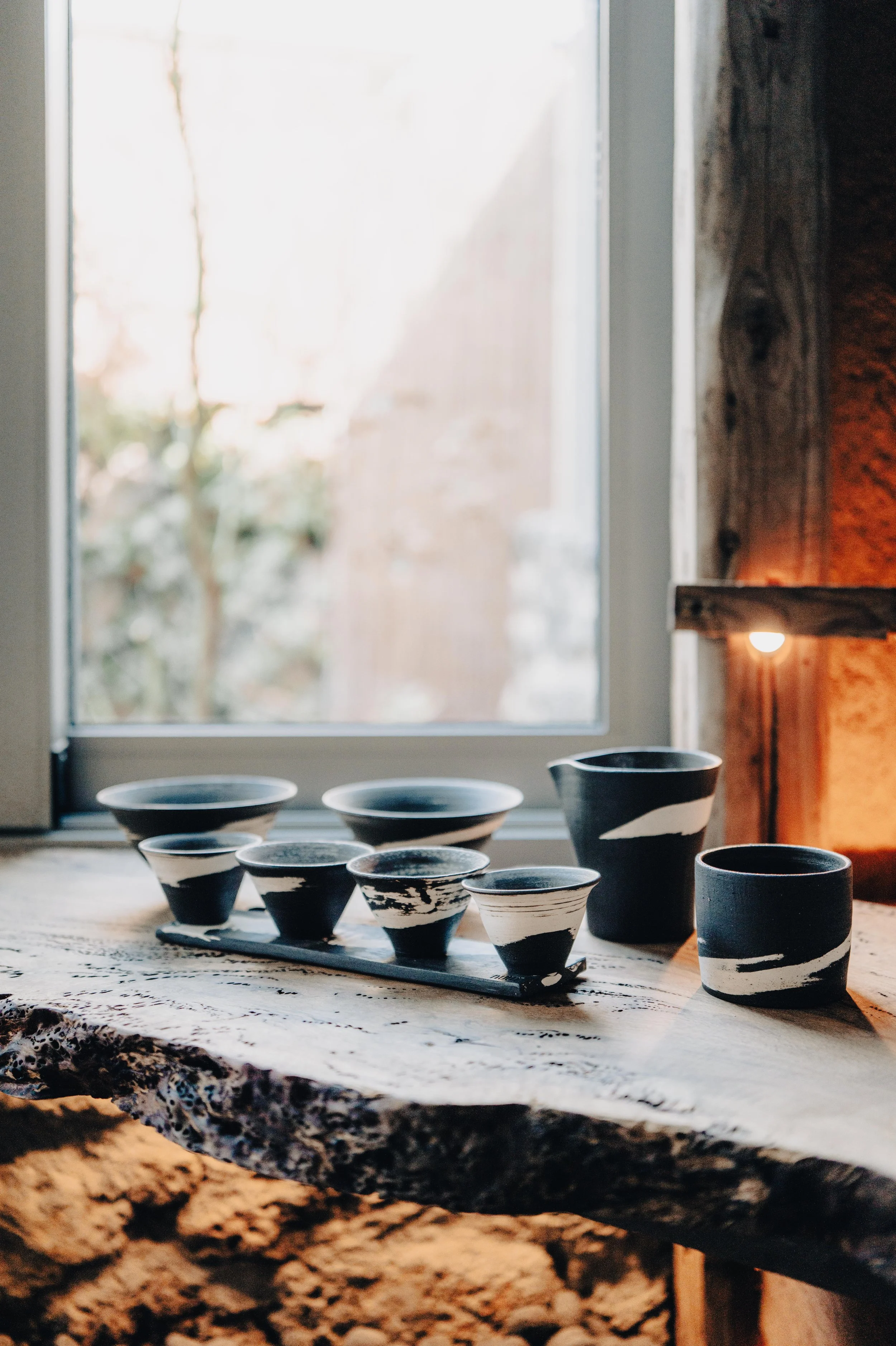 A collection of black and white ceramic cups and bowls on a rustic wooden table near a window.