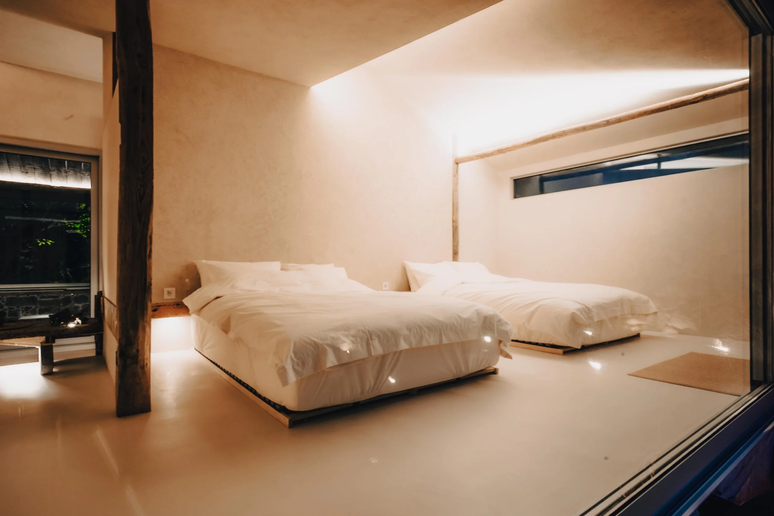 NUUT Aewol 눗애월 A minimalist bedroom with two single beds, white bedding, and warm ambient lighting. Large window on the right side, and a small window on the left. Wooden beams and light-colored walls and floors.