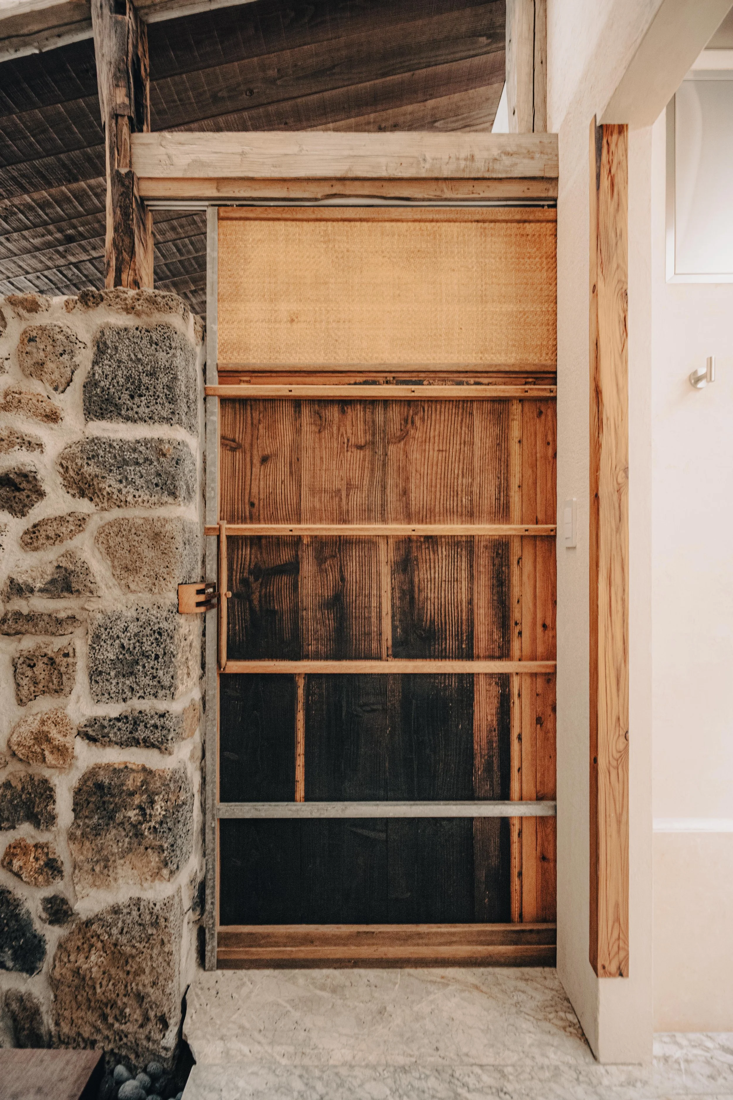 NUUT Aewol 눗애월 Interior view of a wooden sliding door with a stone wall on the left side, part of a rustic room or house.