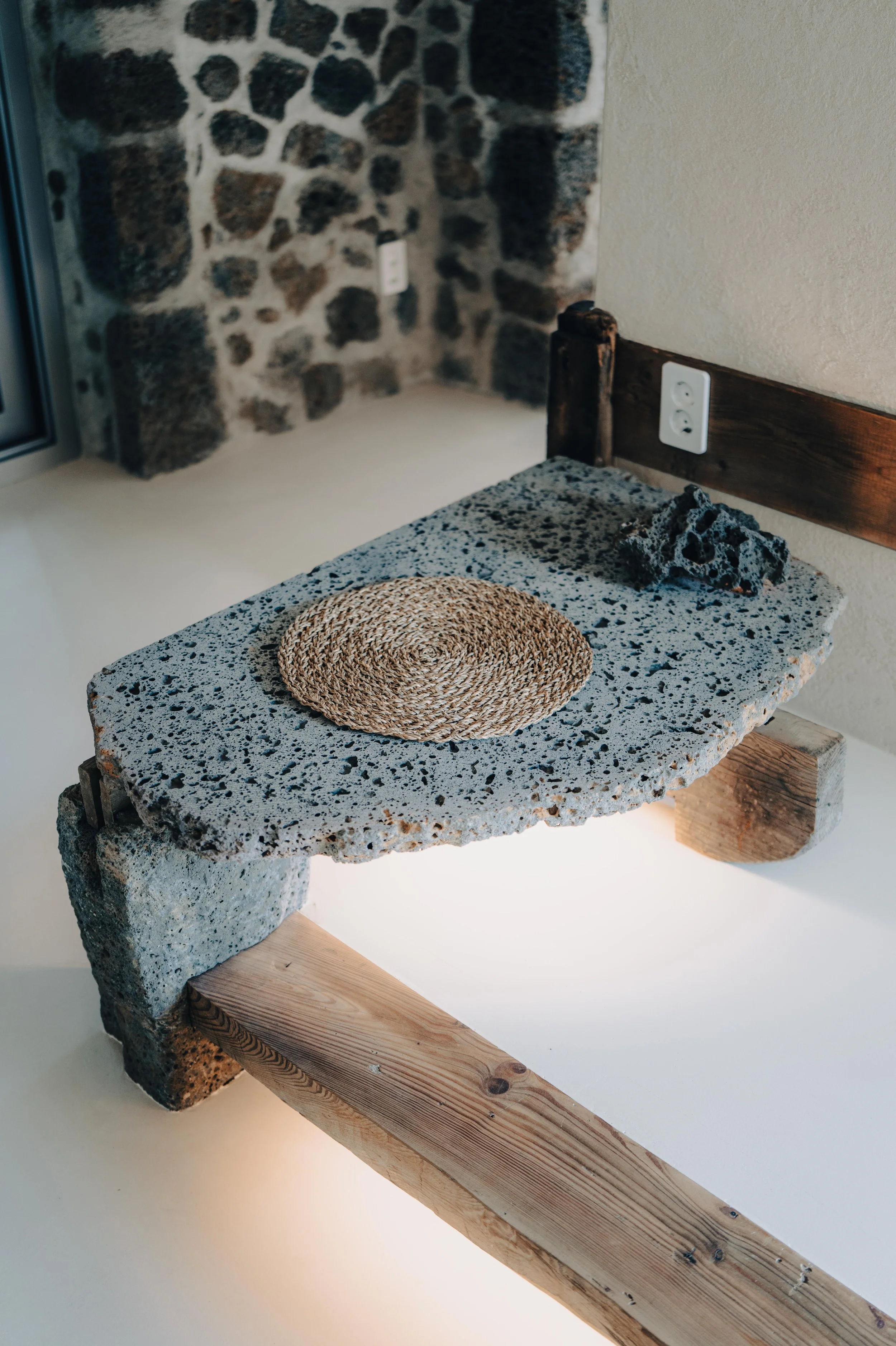 NUUT Aewol 눗애월 A rustic side table made of natural stone and wood pieces with a woven coaster on top, set against a background of a stone wall and a beige wall with an electrical outlet.