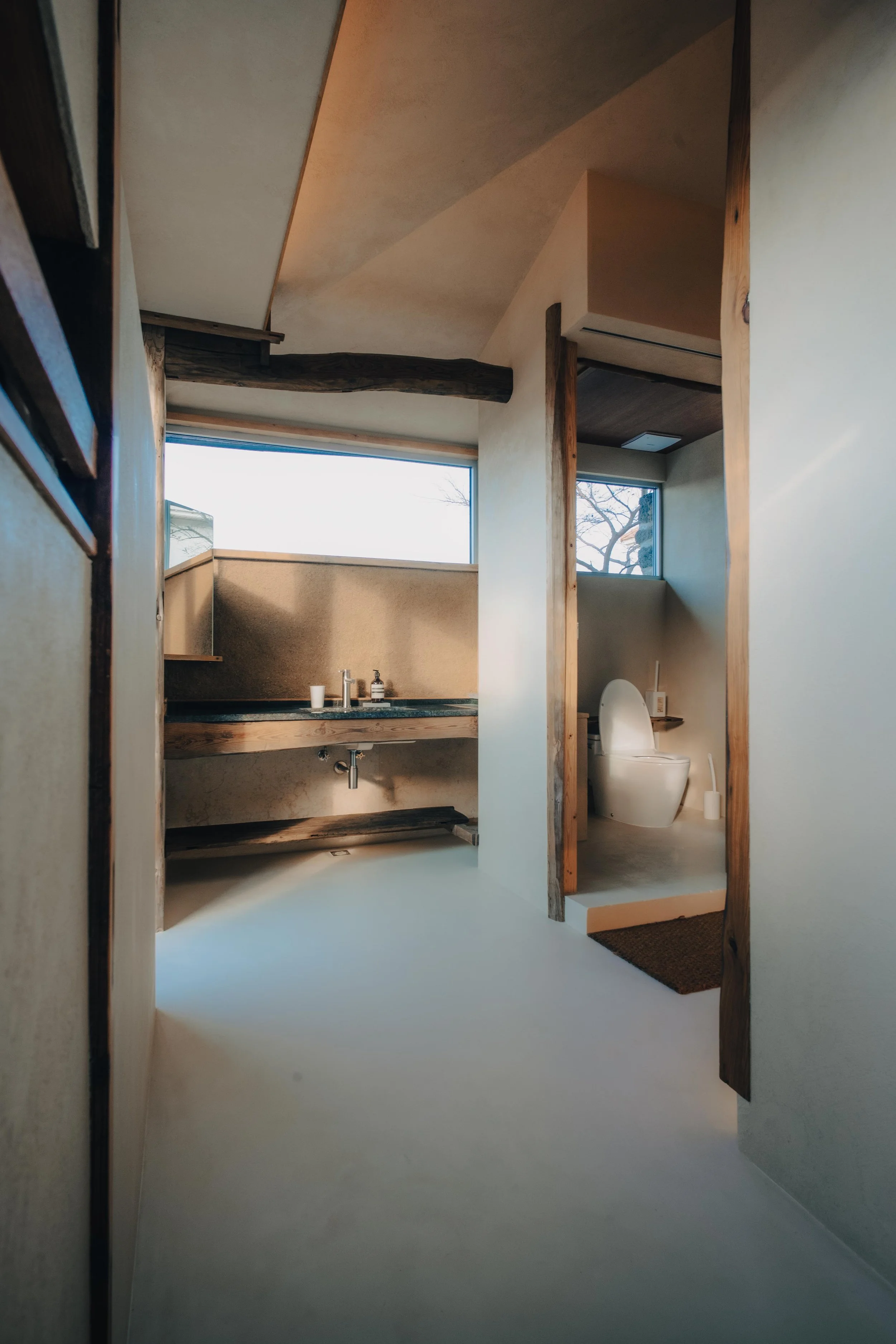 NUUT Aewol 눗애월 Modern bathroom with natural wood accents, a large window, a sink with a dark stone countertop, and a separate toilet area with a small window.