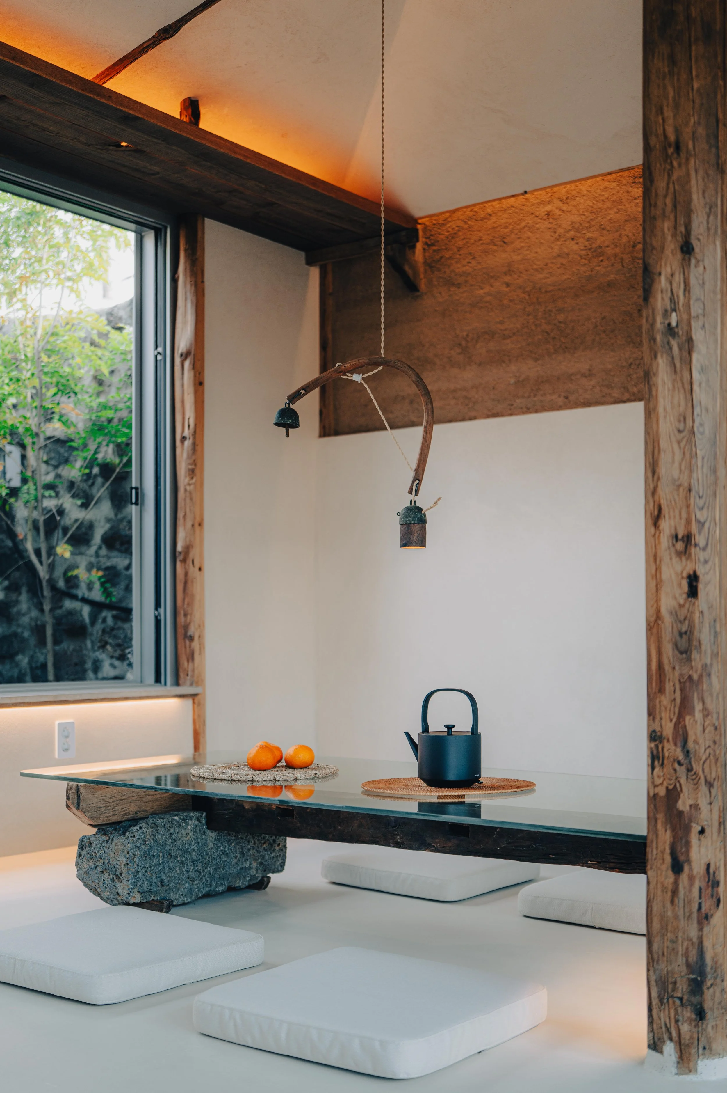 NUUT Aewol 눗애월 A minimalist indoor space with a low wooden table supported by a large stone, surrounded by white cushions on the floor, with a black teapot and oranges on the table, a window showing greenery, and warm ceiling lighting.