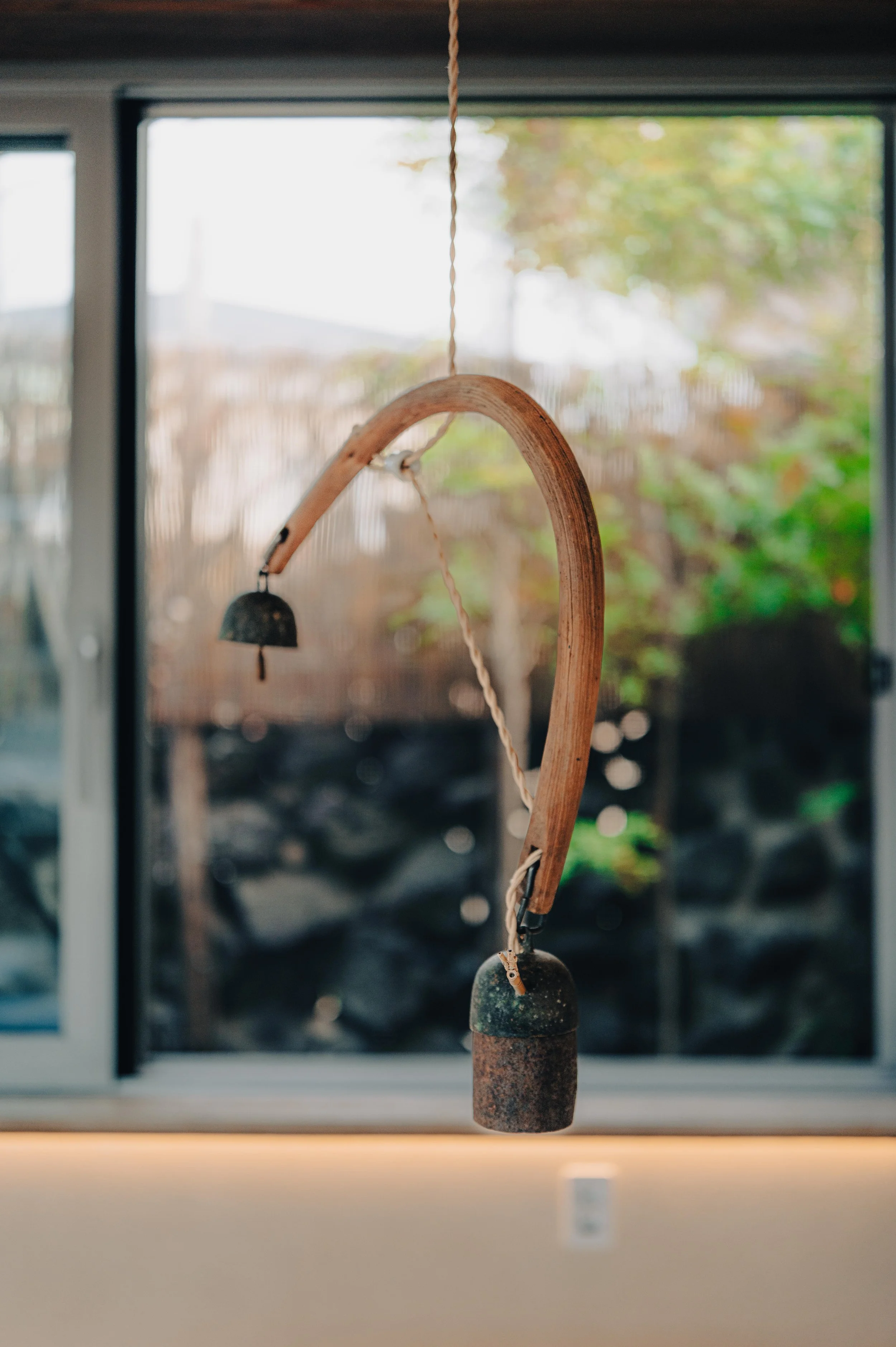 NUUT Aewol 눗애월 A rustic bell mounted on a wooden arch with a rope hanging from it, positioned in front of a window with blurred outdoor greenery in the background.