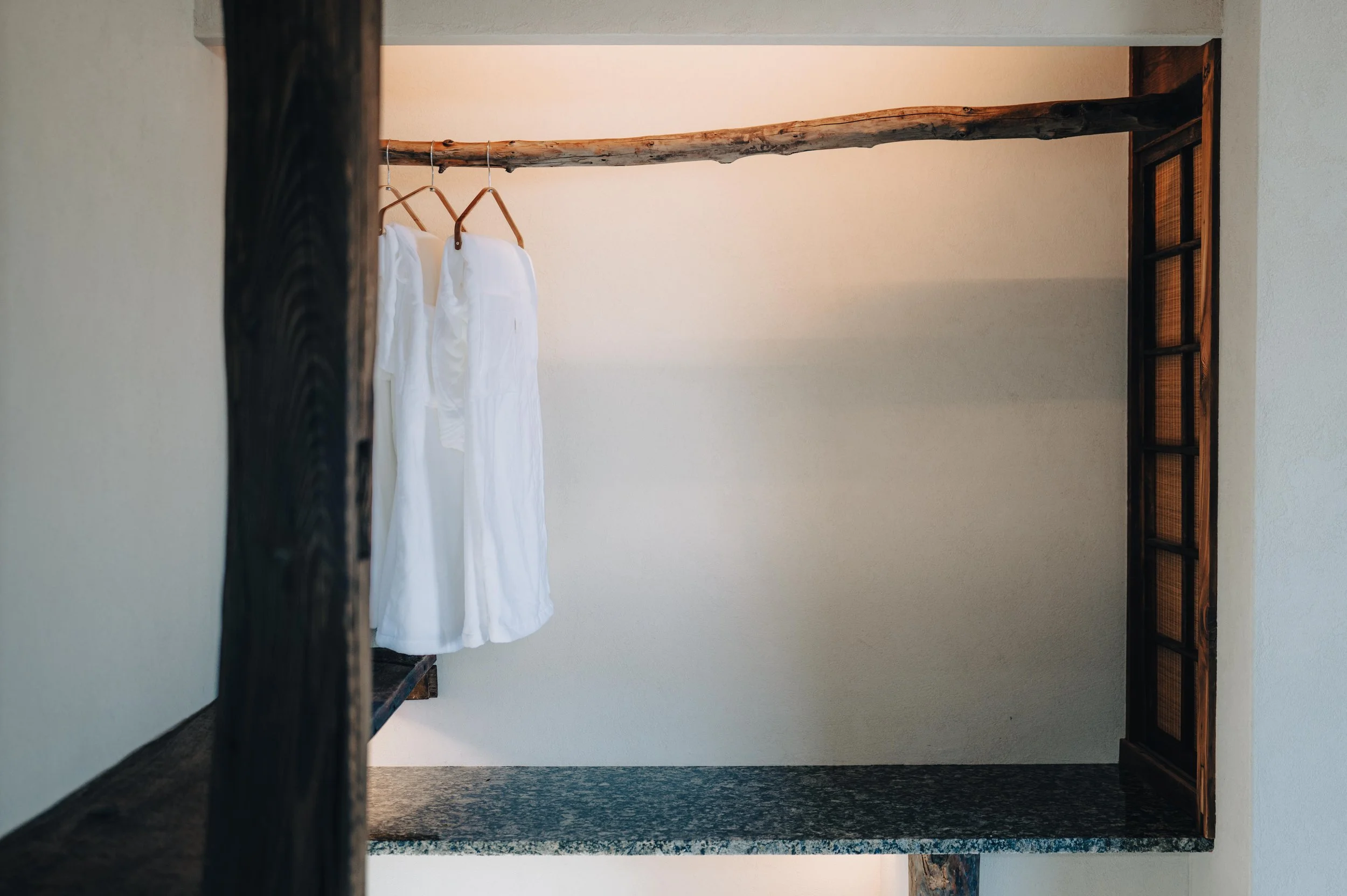 NUUT Aewol 눗애월 View of white clothing or dresses hanging on a wooden rod inside a rustic open wardrobe with dark wood trim and a granite shelf at the bottom.
