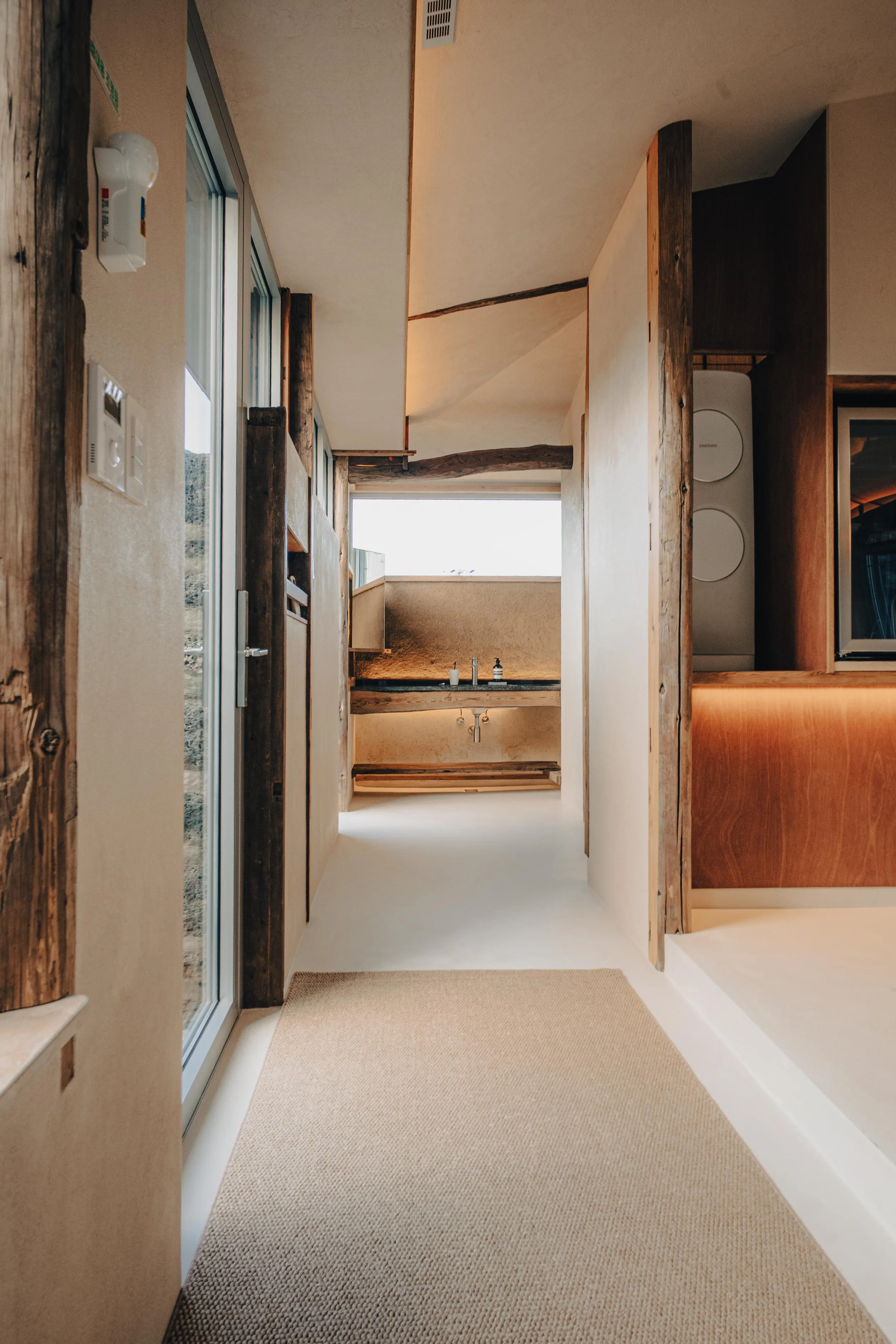NUUT Aewol 눗애월Interior view of a modern home with beige walls, wooden accents, and a carpeted hallway leading to a kitchen area with a sink and large window.