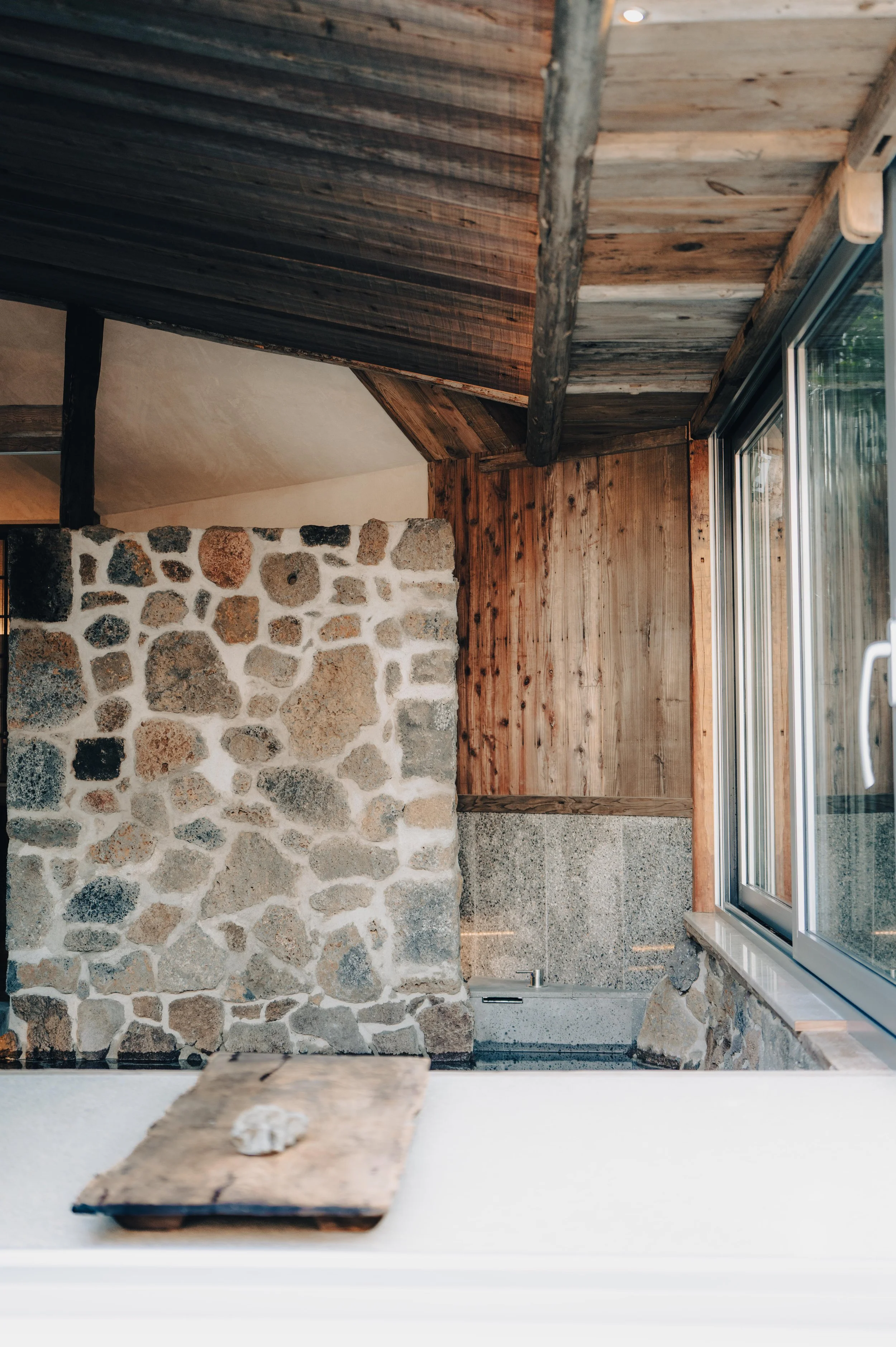Interior of a rustic room with stone, wood-paneled walls, and a large window. A wooden piece with a shell on top is in the foreground.