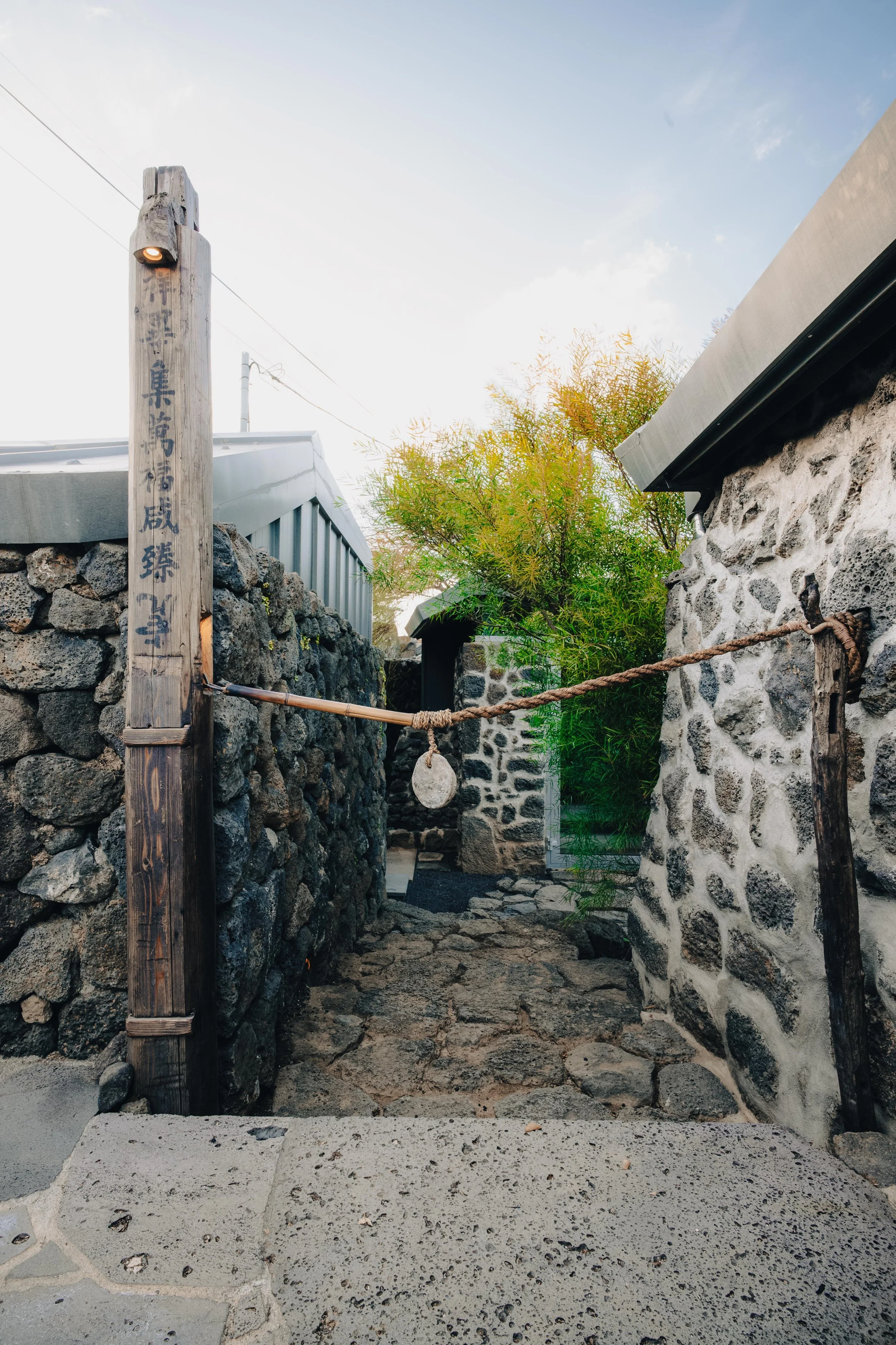 NUUT Aewol 눗애월Stone pathway between traditional stone walls, with a wooden post and rope barrier, and lush green foliage in the background under a clear sky.