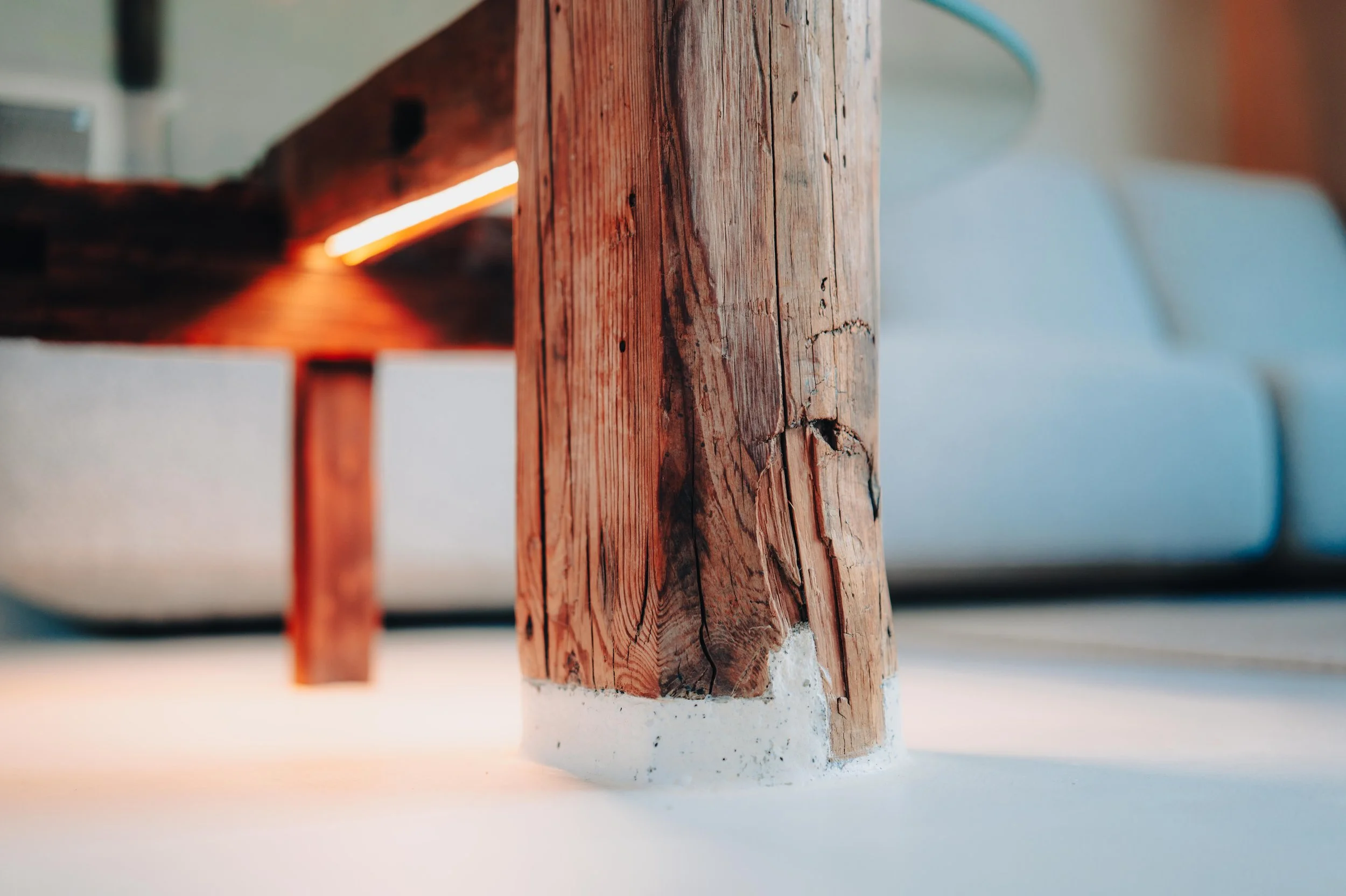 NUUT Aewol 눗애월 Close-up of a wooden table leg with visible grain and cracks, supported on a white concrete base, with a blurred background of a white sofa and part of a room.