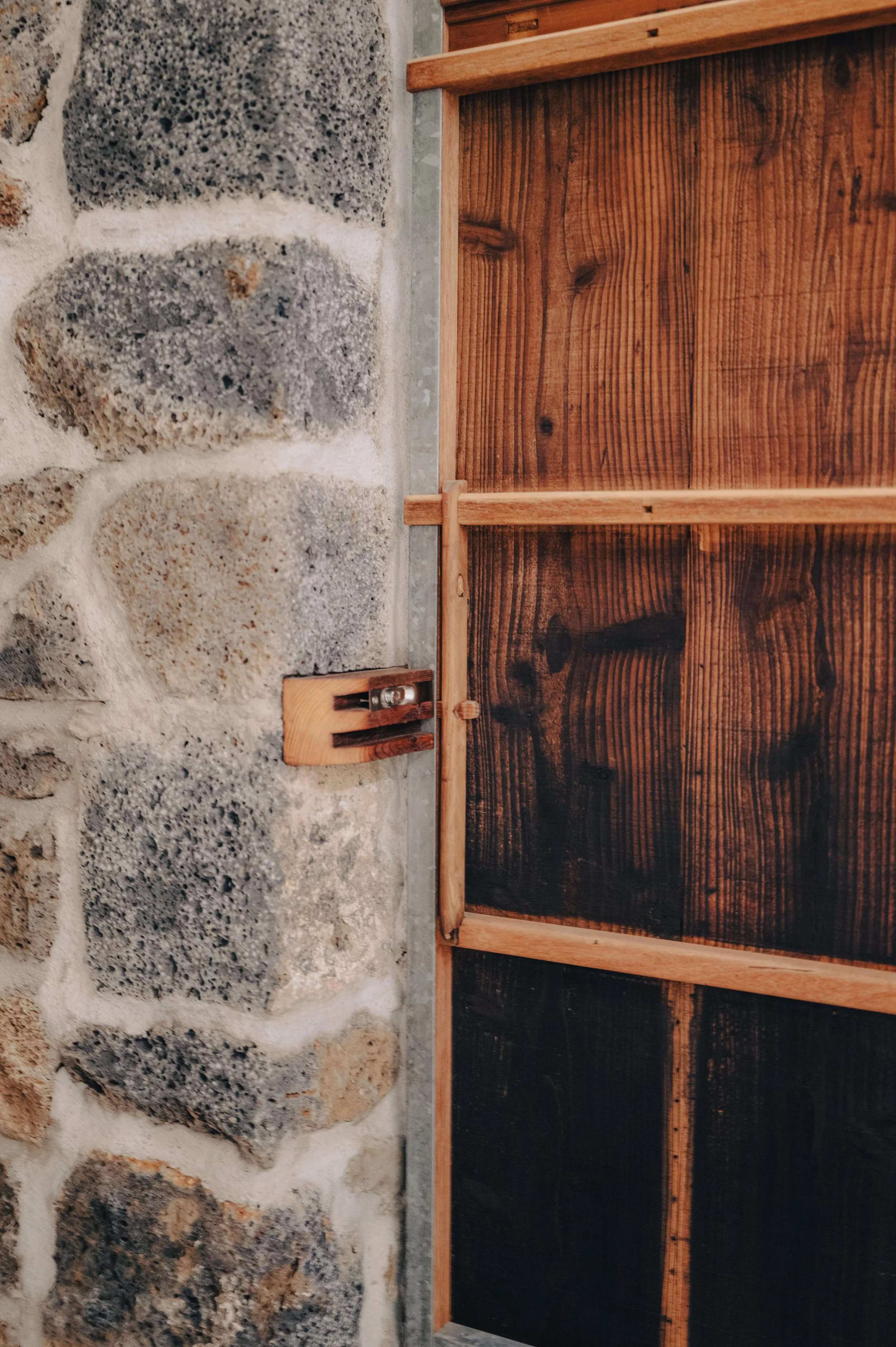 NUUT Aewol 눗애월 Close-up of a stone wall with shades of gray and beige, alongside a wooden structure with vertical and horizontal wooden panels with a stained finish, and a small wooden latch or lock.
