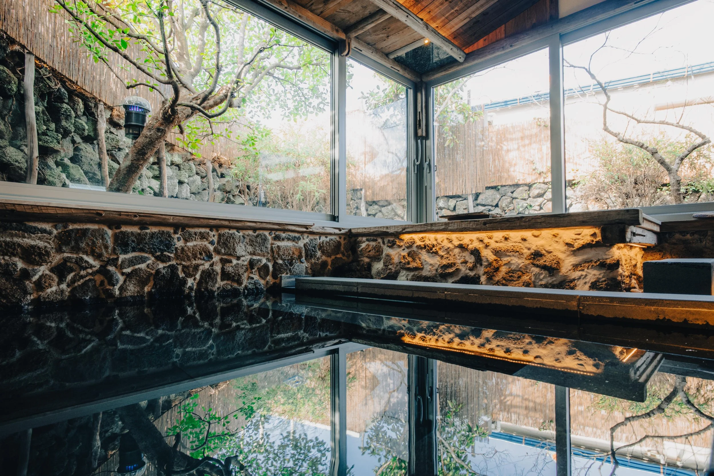 NUUT Aewol 눗애월 View of a small outdoor hot spring or soaking tub with a reflection of nearby trees and sky, surrounded by stone walls and large windows.