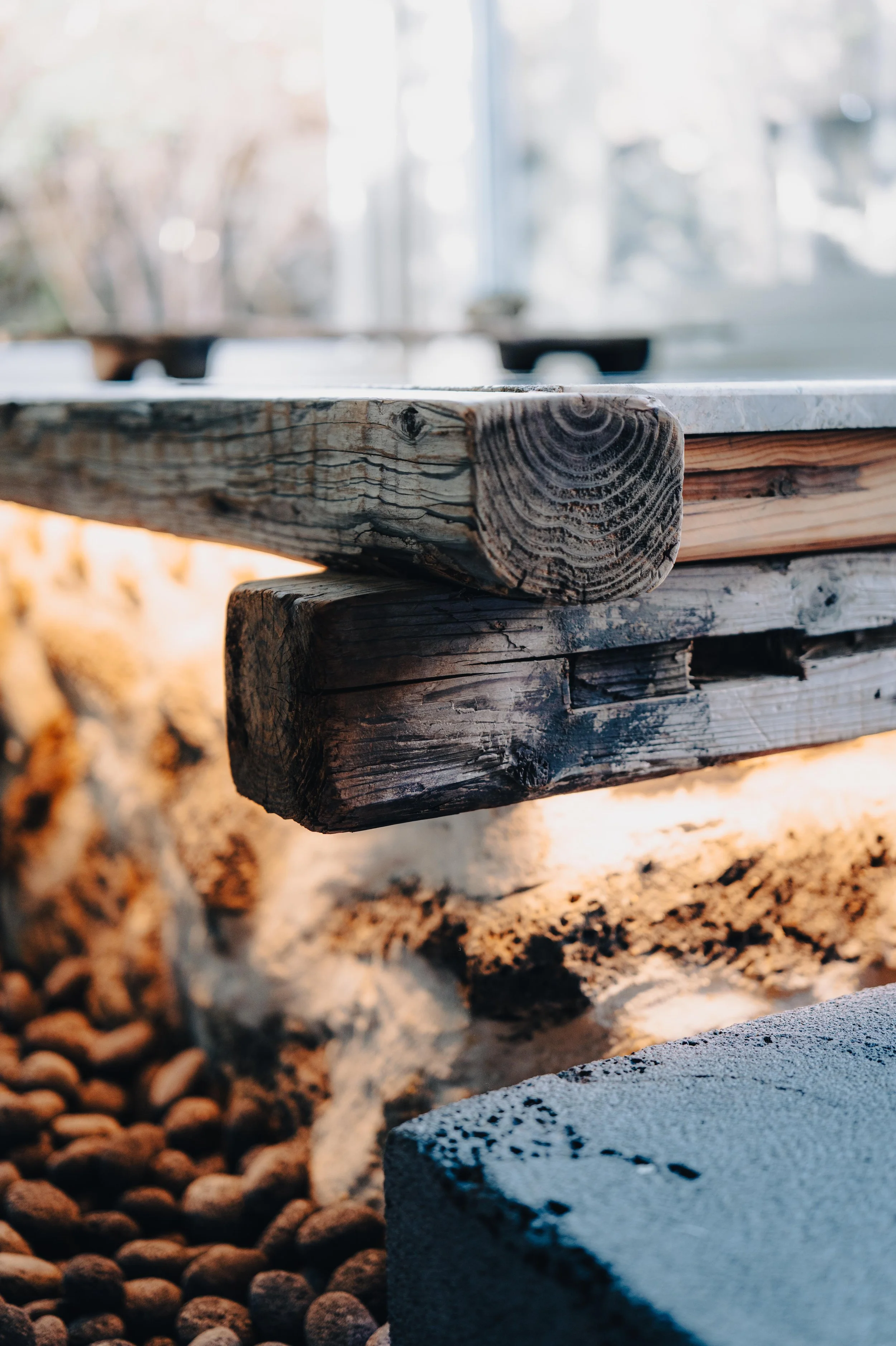 Close-up view of a rustic wooden table with a marble top, supported by a blackened wooden beam, set on a bed of small pebbles with soft natural light in the background.