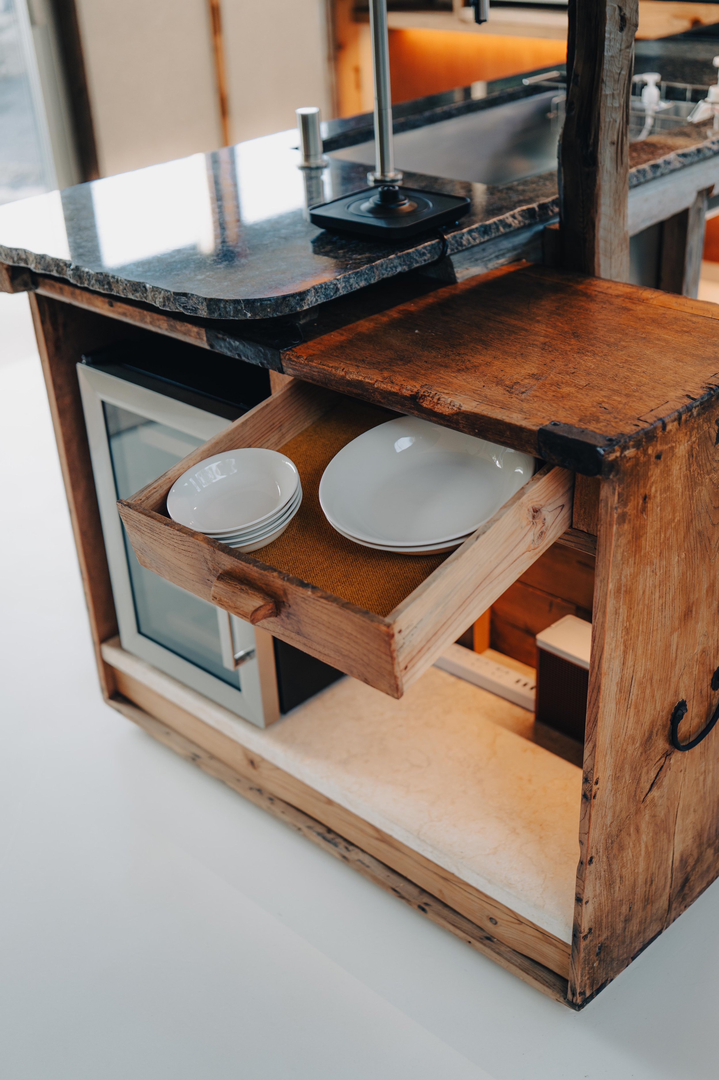 NUUT Aewol 눗애월 A rustic wooden kitchen cart with a drawer holding white dishes and a small mini fridge underneath, a granite countertop, some baking tools, and a wooden shelf beside it.