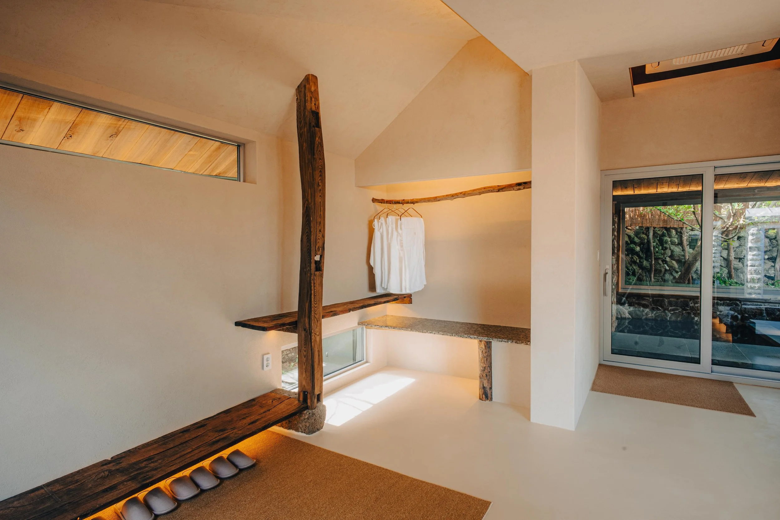 NUUT Aewol 눗애월 Modern minimalist interior with wooden accents, a sliding glass door leading outside, and a small hanging white robe.