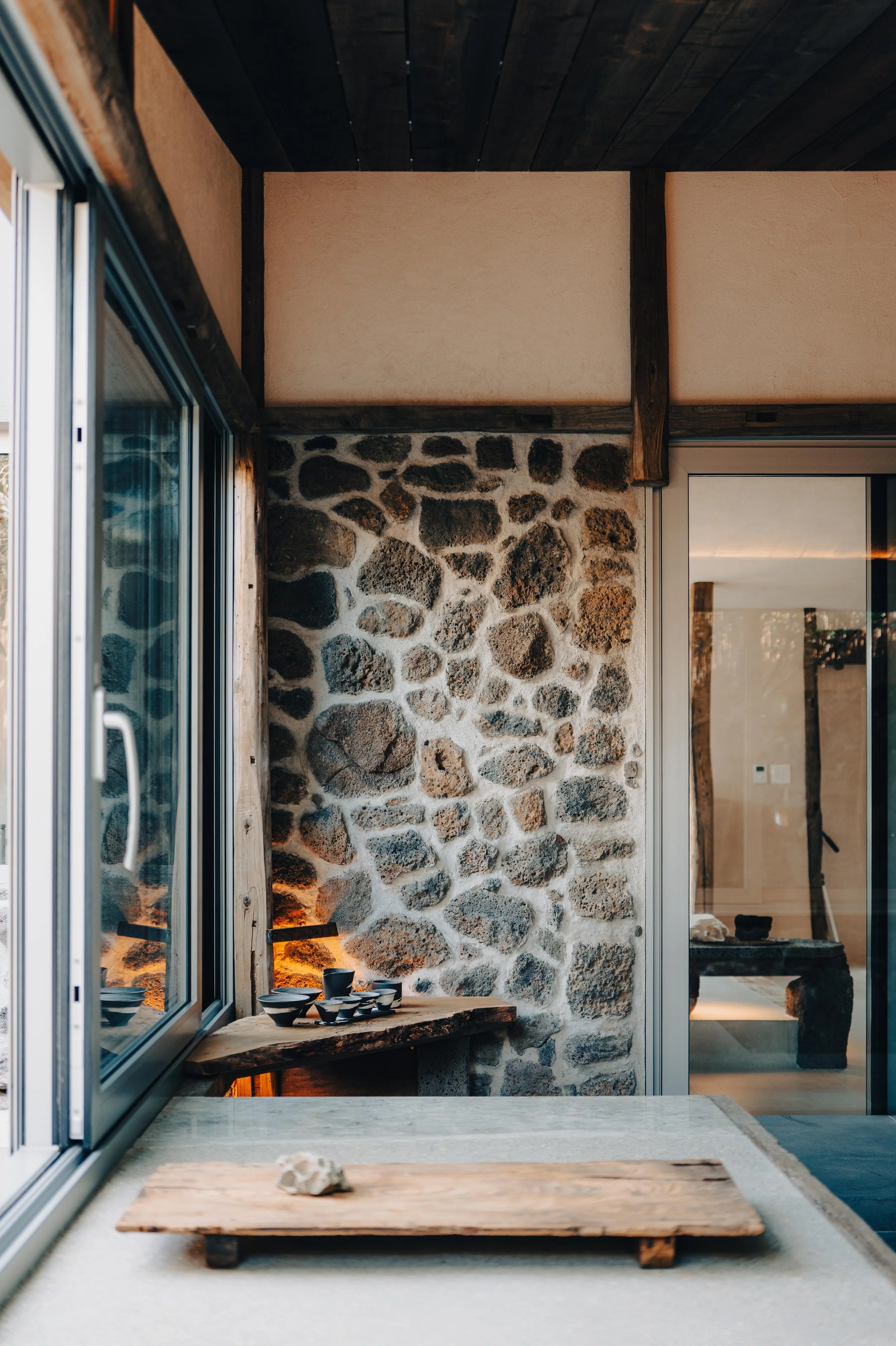 Interior view of a rustic room with stone wall, wooden ceiling, and glass sliding doors, showcasing decorative black bowls and a piece of wood with a small sculpture on a countertop.